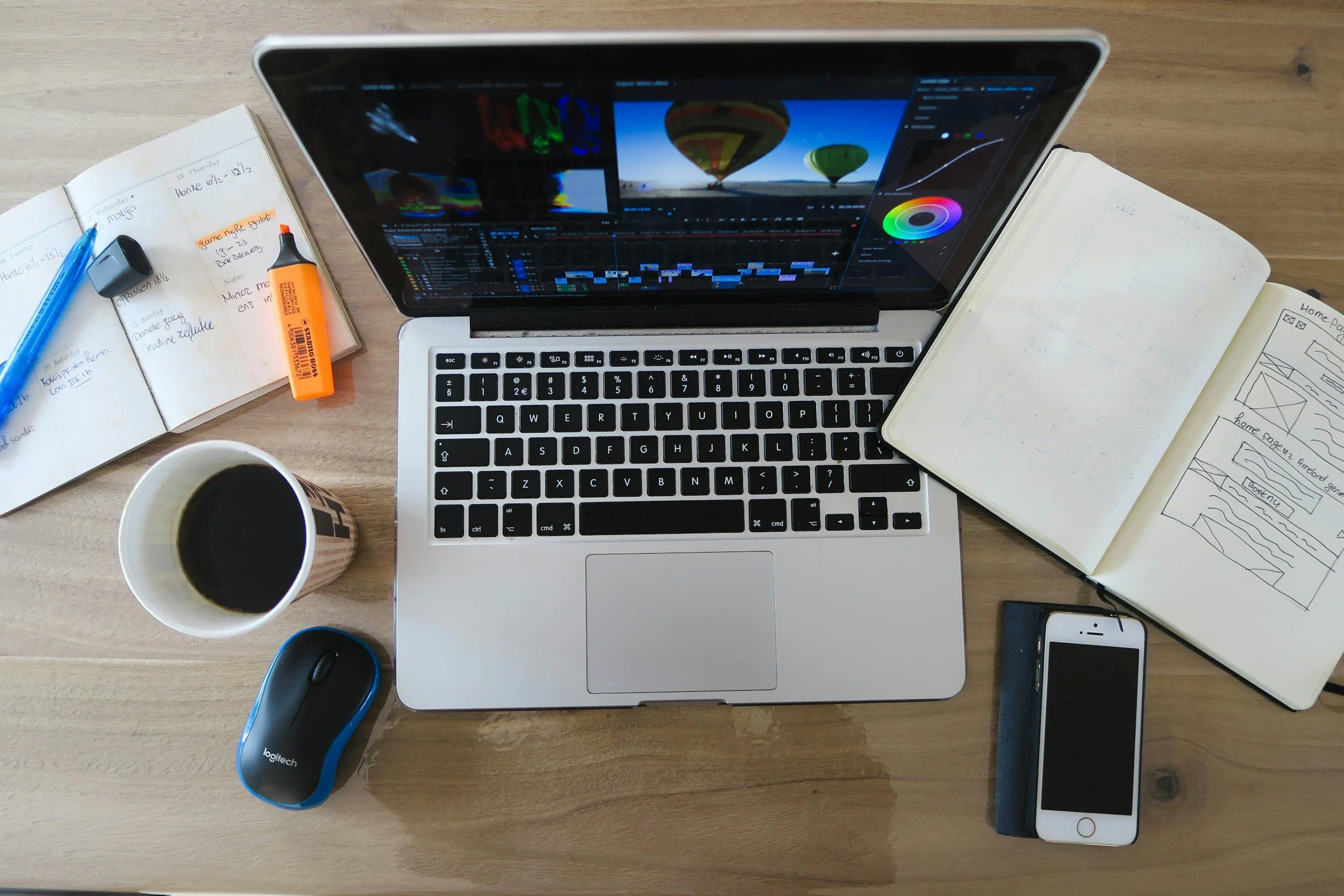 Top-down view of a wooden desk with a silver laptop displaying video editing software, a cup of coffee in a paper cup, a wireless mouse, an iPhone in a black case, an open notebook with sketches, a planner with handwritten notes, a blue pen, a highlighter, and an external hard drive.