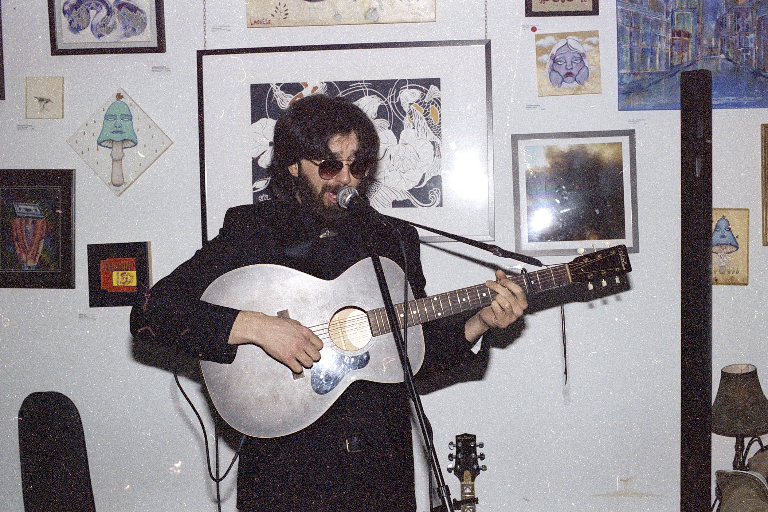 A man with dark hair, sunglasses, and a beard playing an acoustic guitar and singing into a microphone in front of a wall decorated with various framed artworks.