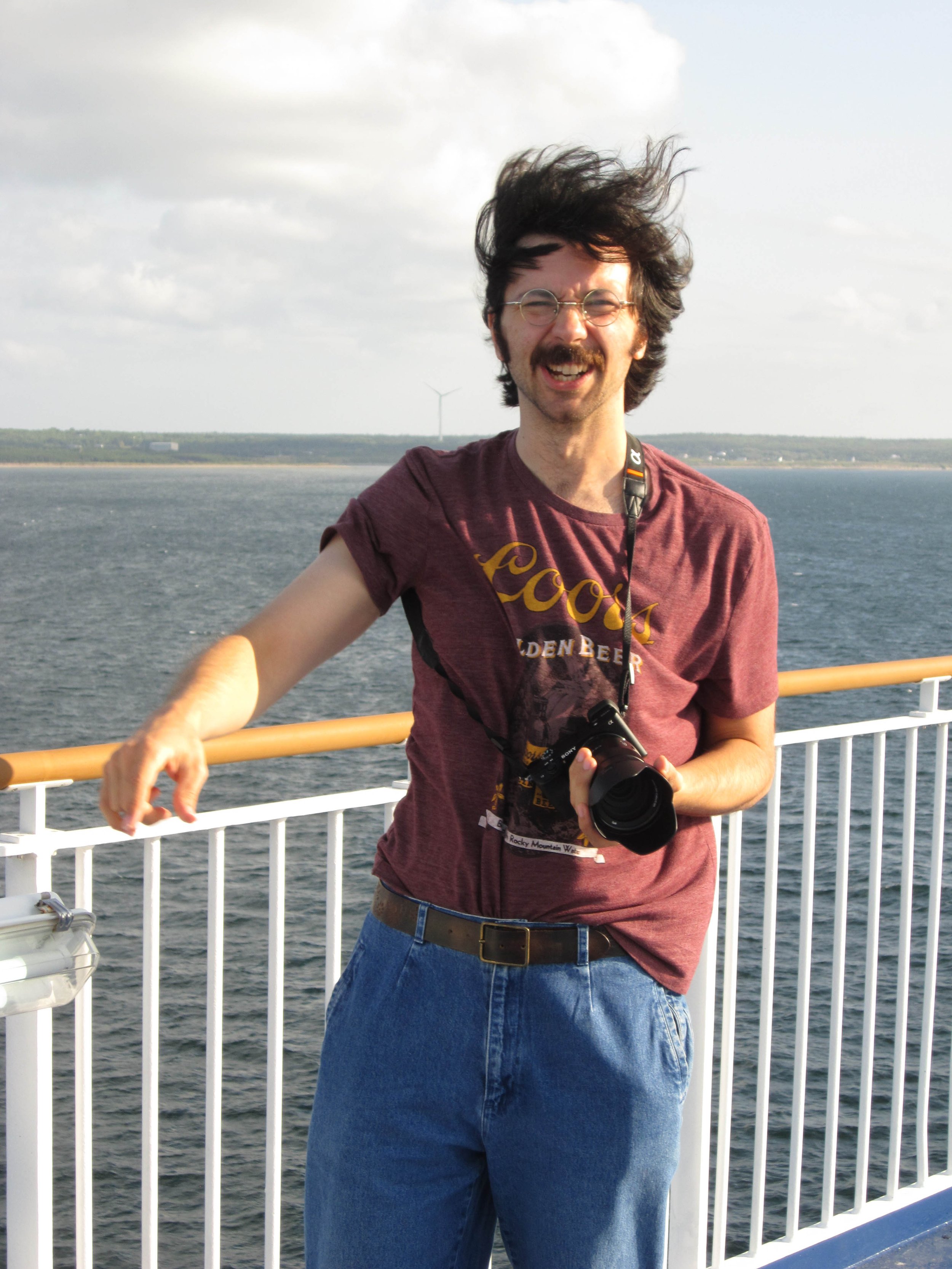 A man standing on a ship deck, smiling, with a camera hanging around his neck, overlooking the water with a wind turbine in the background.