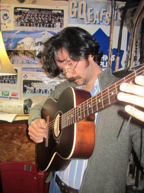 A man with glasses playing an acoustic guitar in a room decorated with sports memorabilia and posters.