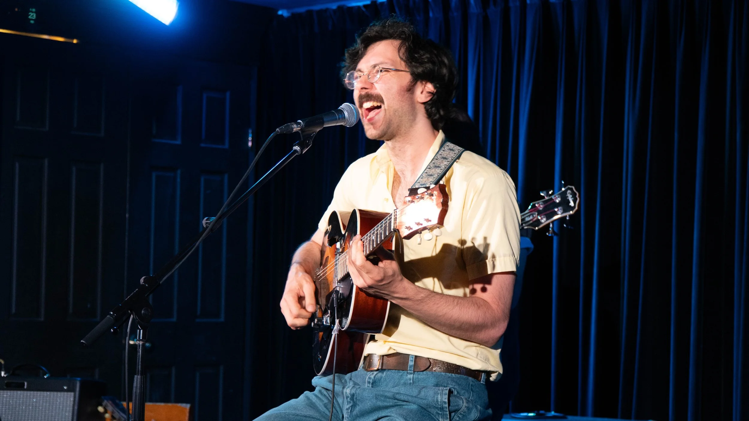 A man with glasses and dark hair, wearing a light yellow shirt, sings into a microphone while playing an acoustic guitar on stage with a dark curtain backdrop.