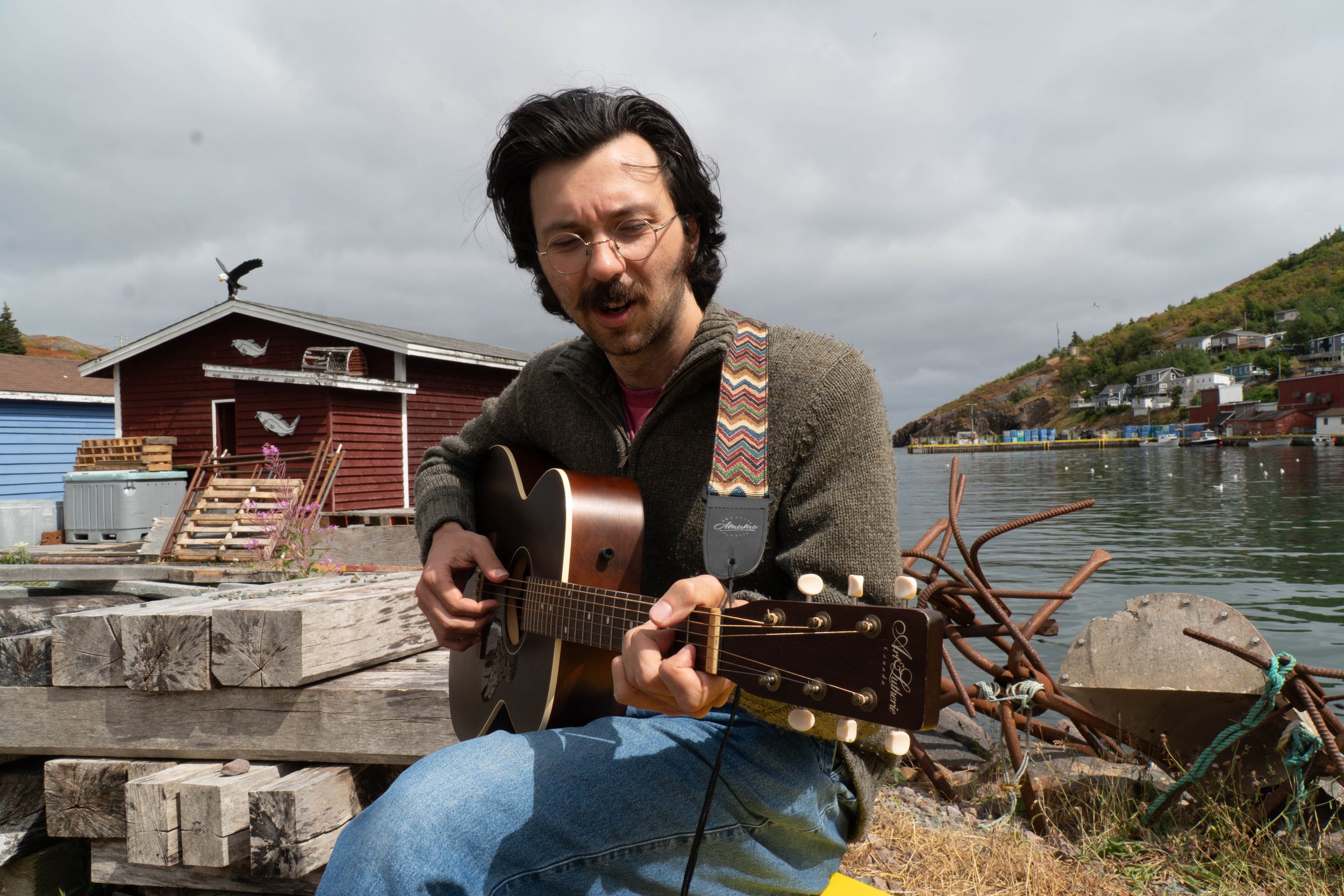 Man playing guitar outdoors near waterfront with colorful houses on hillside in the background