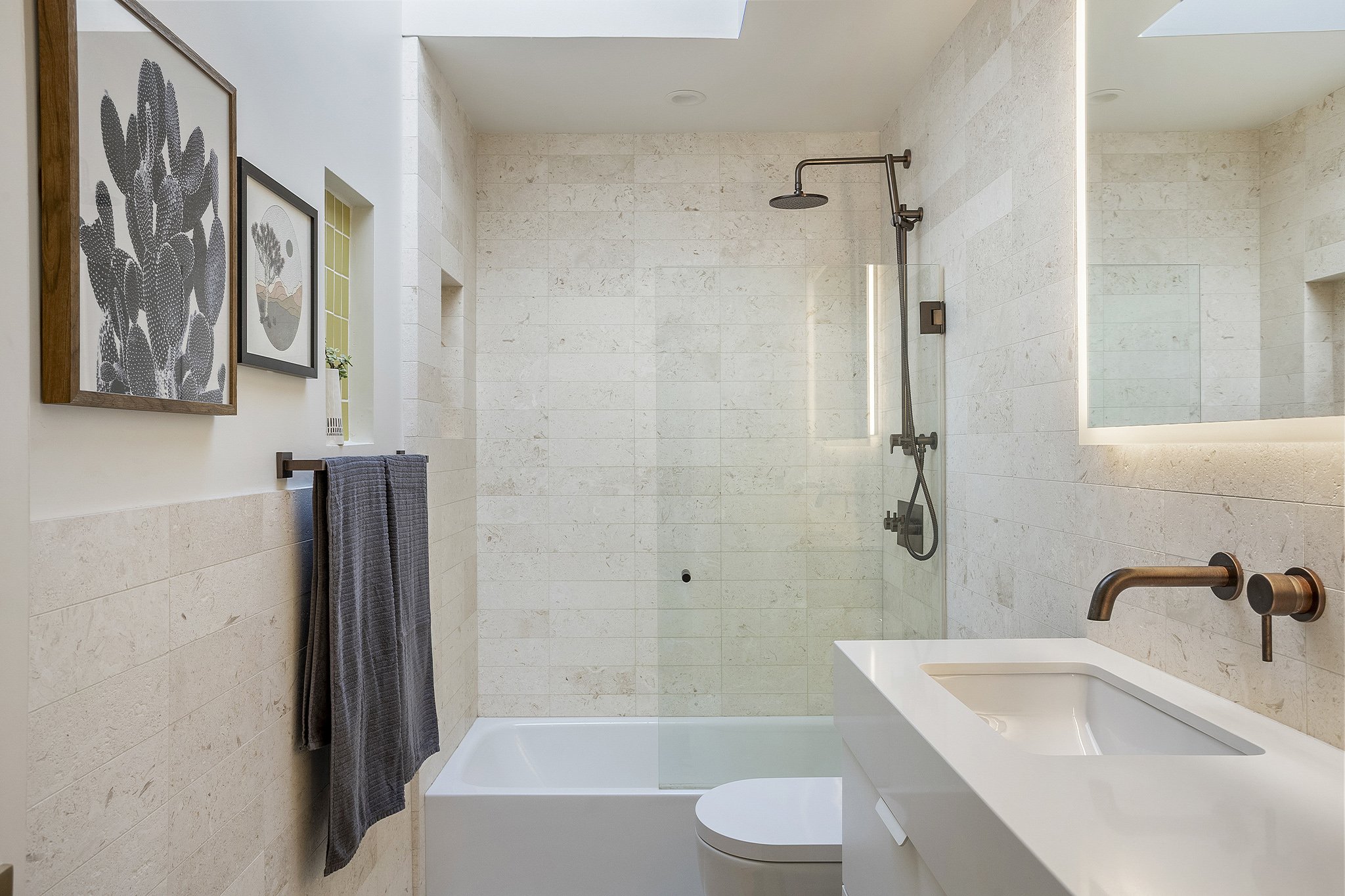 Modern bathroom with a walk-in shower, a white toilet, a white vanity with a copper faucet, and wall art including a cactus and landscape paintings.