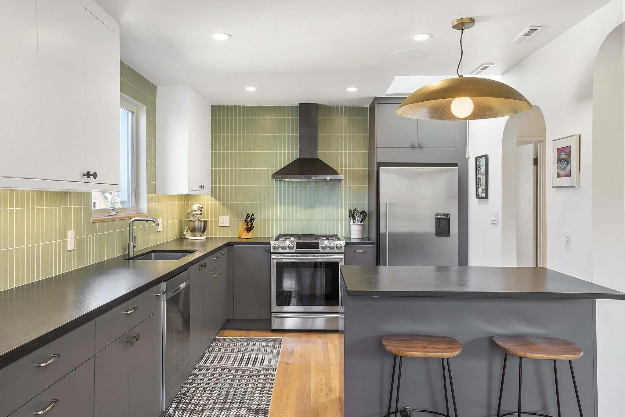 Modern kitchen with green tiled backsplash, gray cabinets, stainless steel stove, fridge, and black countertops, with two wooden bar stools and a large hanging gold light fixture.
