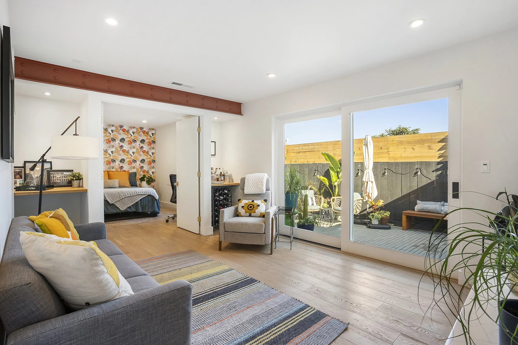 Living room with sliding glass door leading to a patio, a gray sofa with pillows, a gray armchair, a colorful rug, and a bedroom visible through an open doorway with a patterned accent wall.