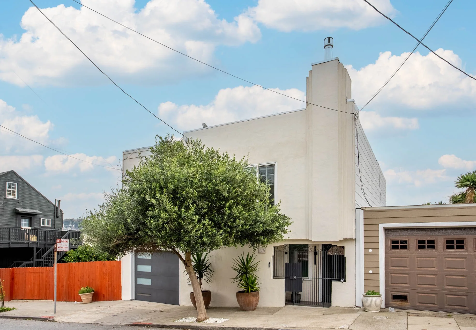 A modern white multi-story house with a black gate, potted plants, and a tree in front, adjacent to a brown garage door, under a blue sky with clouds.