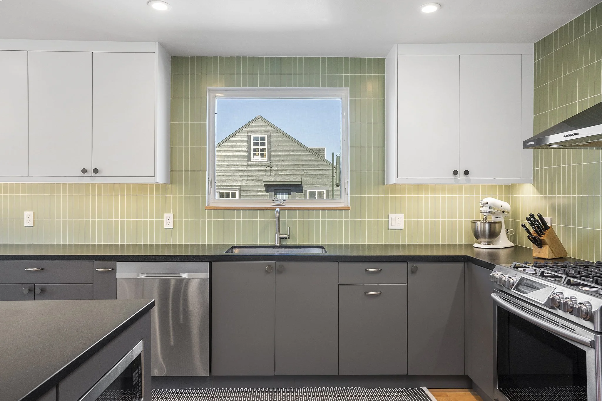 Modern kitchen with white upper cabinets, gray lower cabinets, black countertops, and green tiled backsplash. Contains a window over the sink, stainless steel appliances, and a stand mixer on the countertop.