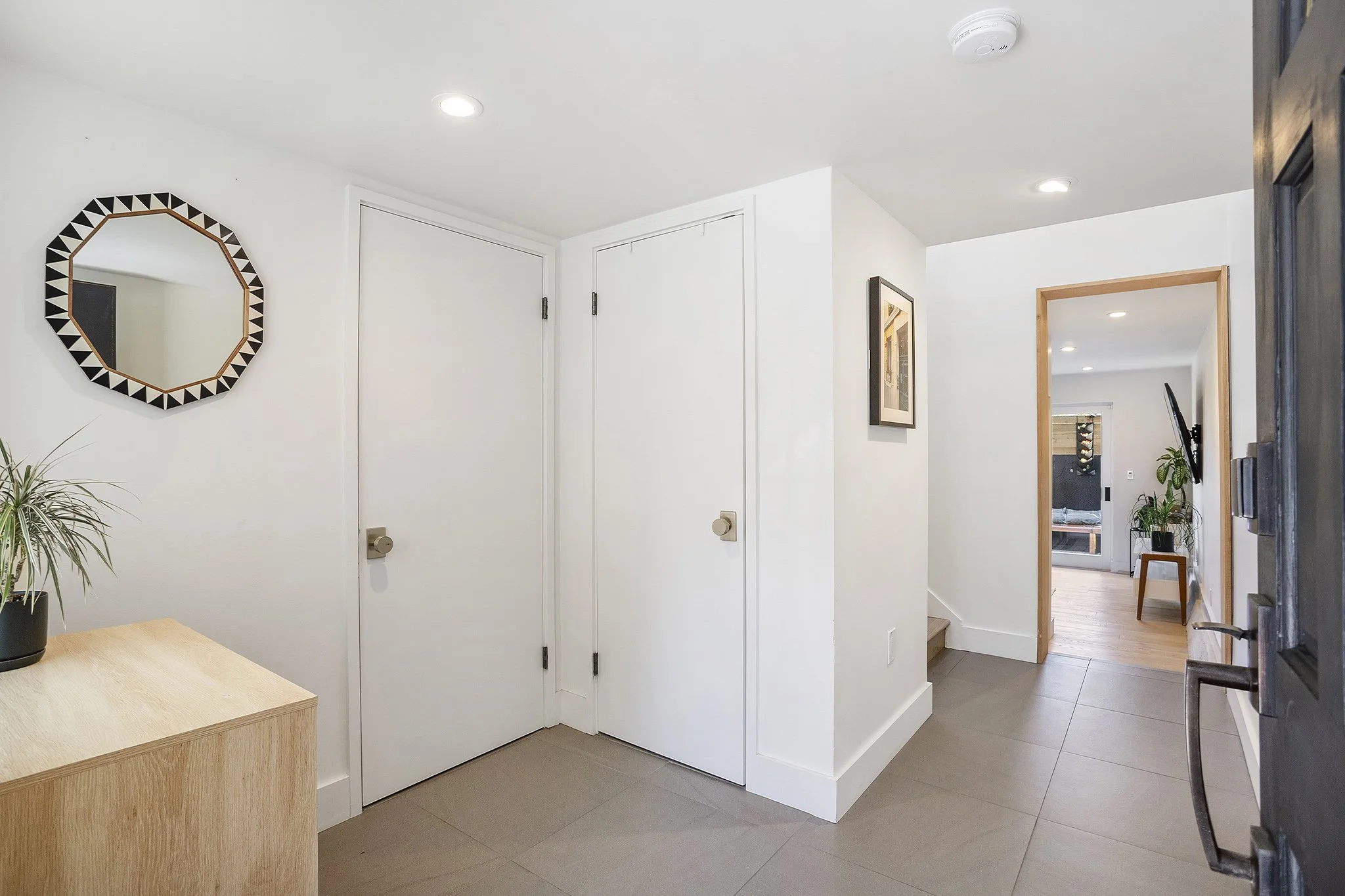 Interior view of a modern home entryway with white walls, wooden accents, a decorative mirror, and potted plants.
