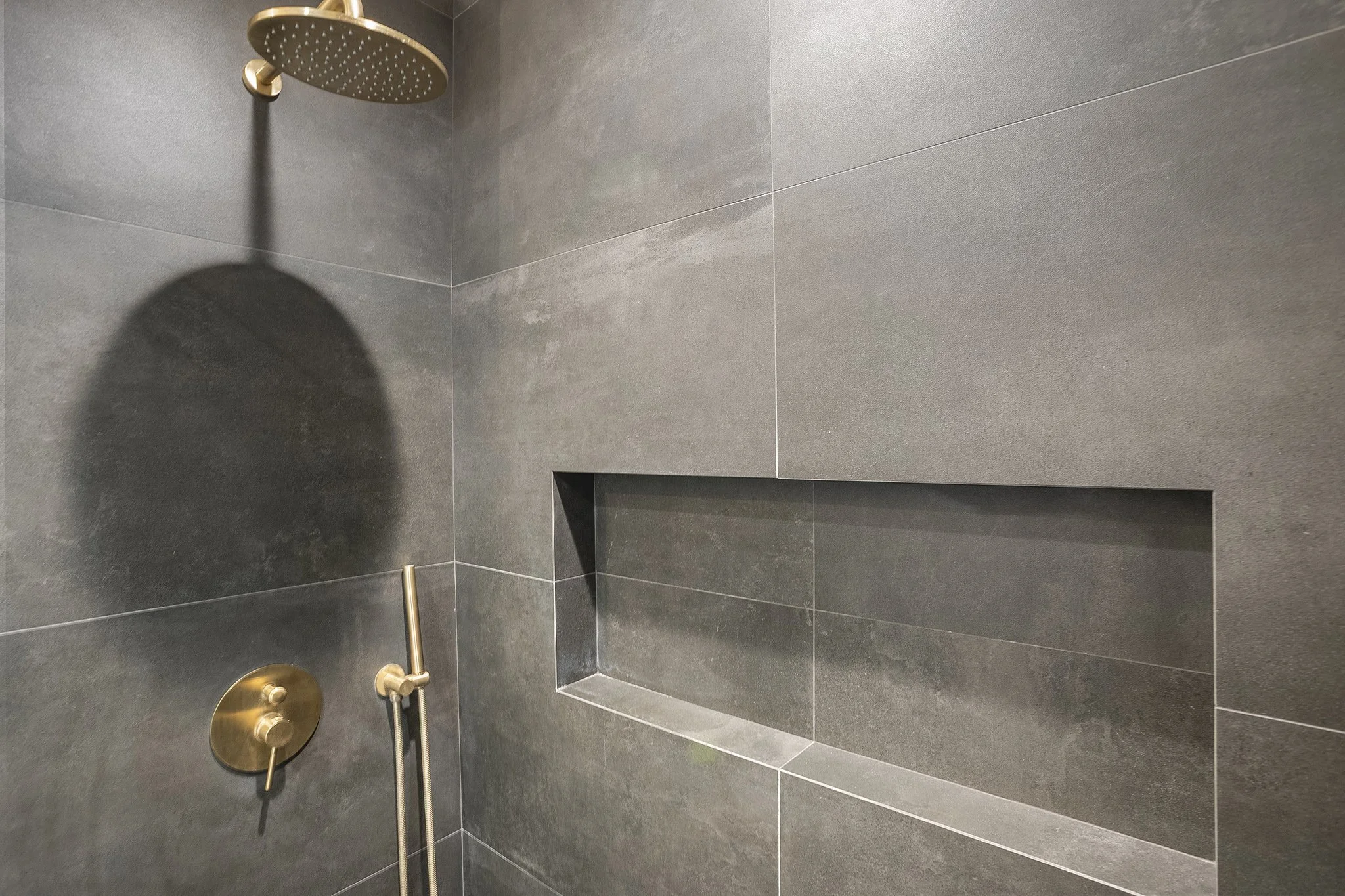 A modern shower with dark gray tiles, a gold showerhead, a gold control valve, and a built-in wall niche.
