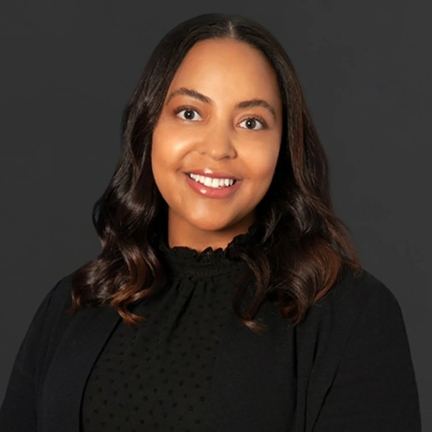A young woman with long wavy hair and a warm smile wearing a black top against a dark gray background.