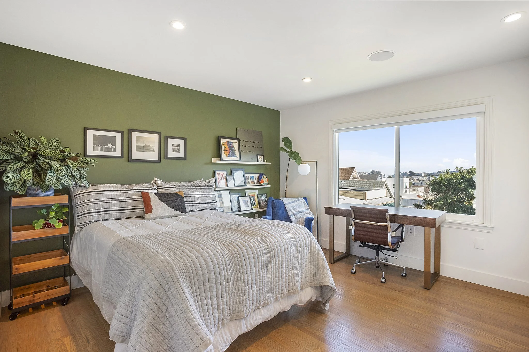 Bedroom with a green accent wall, bed with striped pillows, nightstand with a plant, framed pictures above the bed, a large window with a city view, a small desk with a chair, and a blue armchair with a floor lamp.