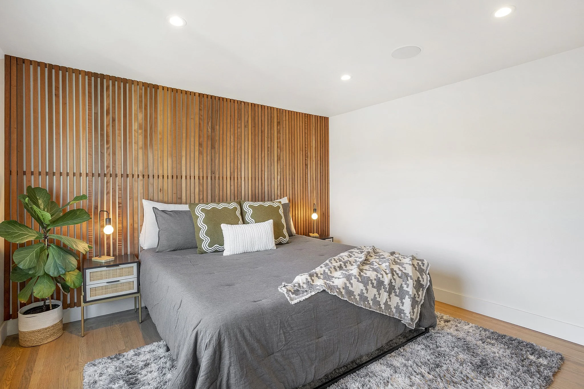 A modern bedroom with a bed against a wooden slat wall, gray bedding, decorative pillows, a fuzzy rug, a large green houseplant, and small bedside tables with lamps.