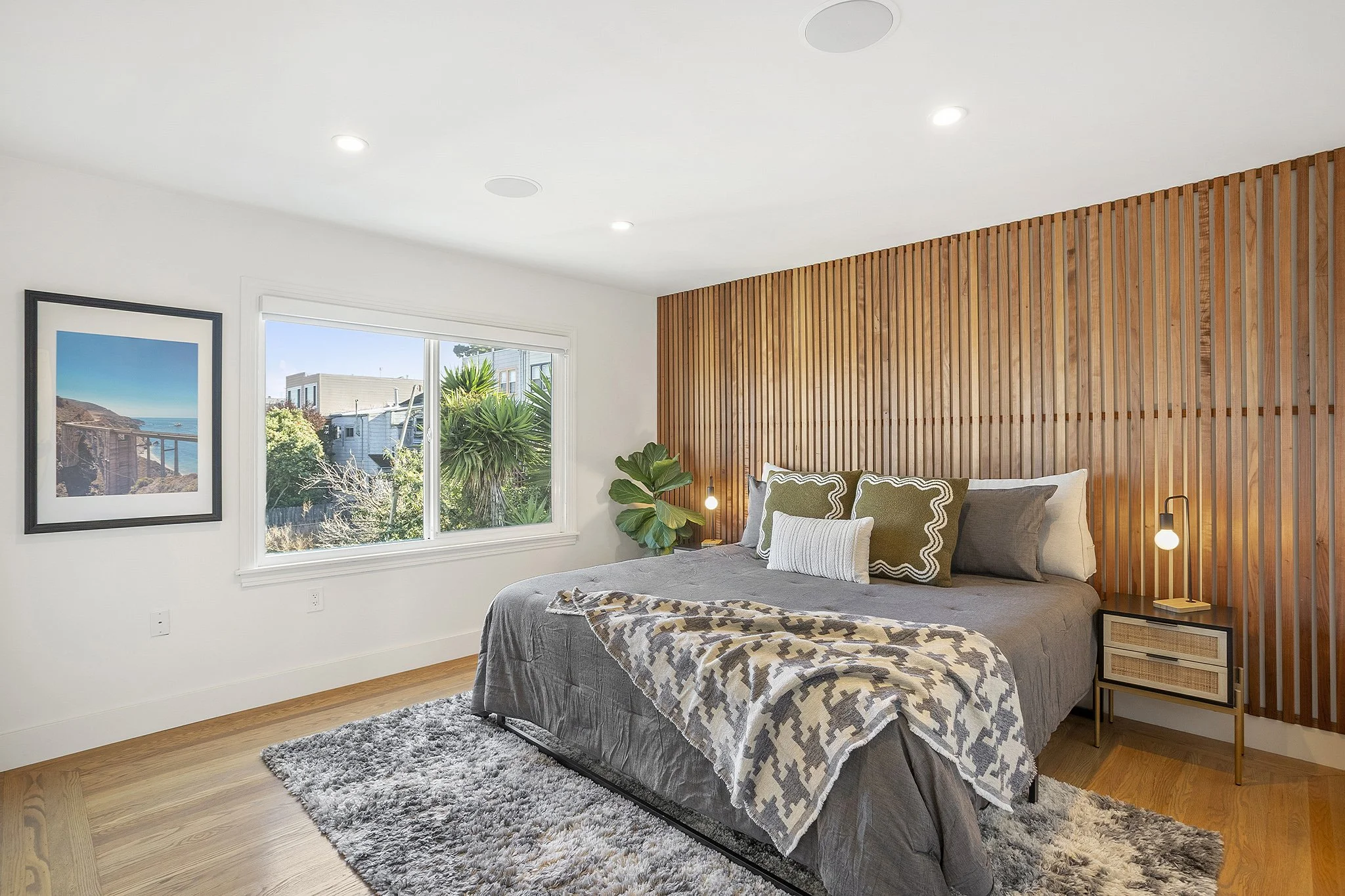 A modern bedroom featuring a bed with gray bedding, decorative pillows, and a blanket. The room has a wooden accent wall behind the bed, a bedside table with a lamp, and a large window with greenery outside. There is a framed landscape photograph on 