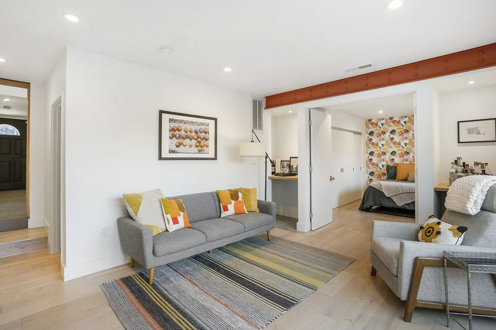 Living room with gray sofa, colorful pillows, framed artwork, room divider, armchair with floral pillow, and entryway with black door.