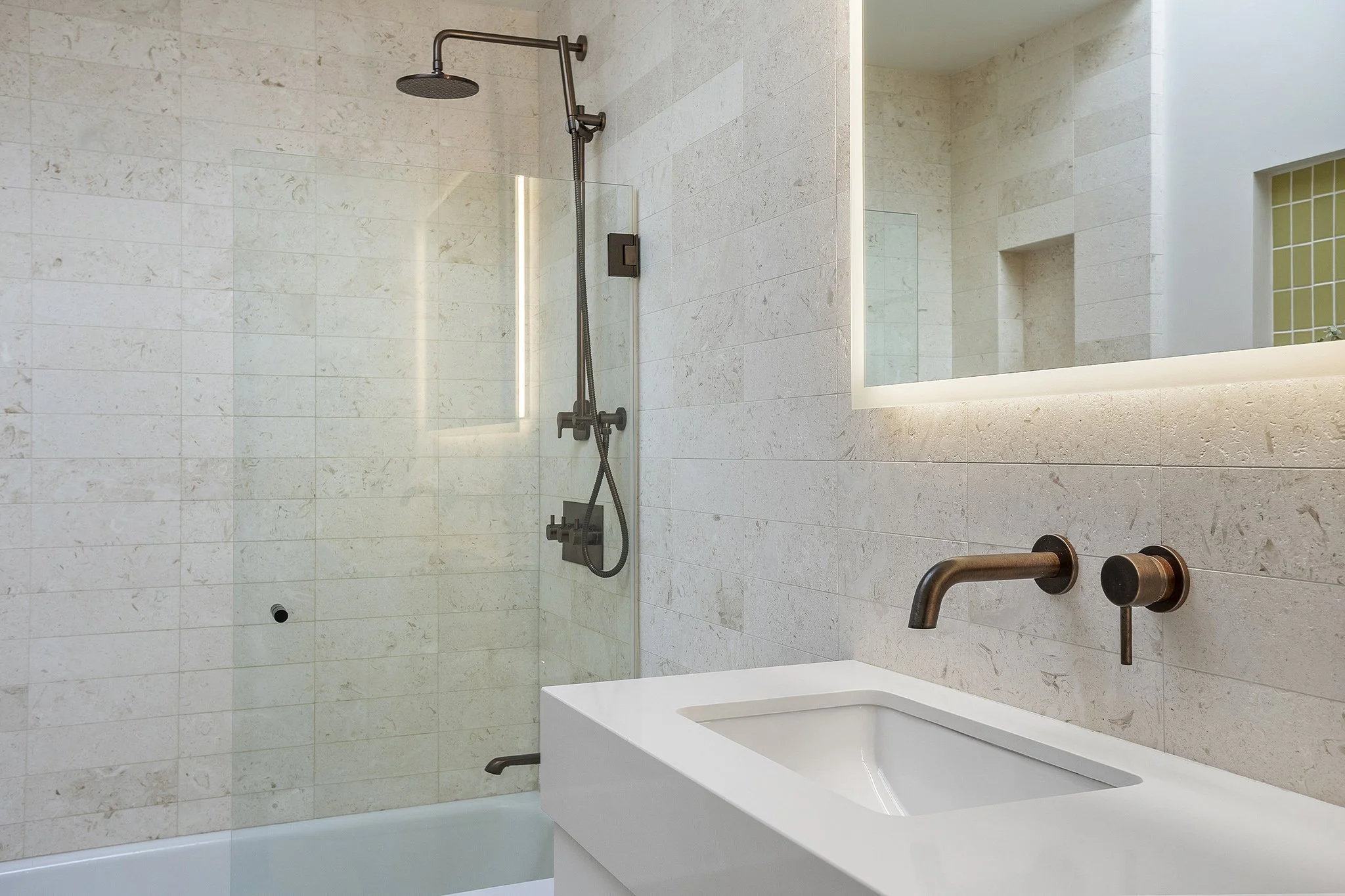 Modern bathroom with beige tiled walls, a walk-in shower with a dark metal showerhead, a white rectangular sink with bronze fixtures, and a backlit mirror.