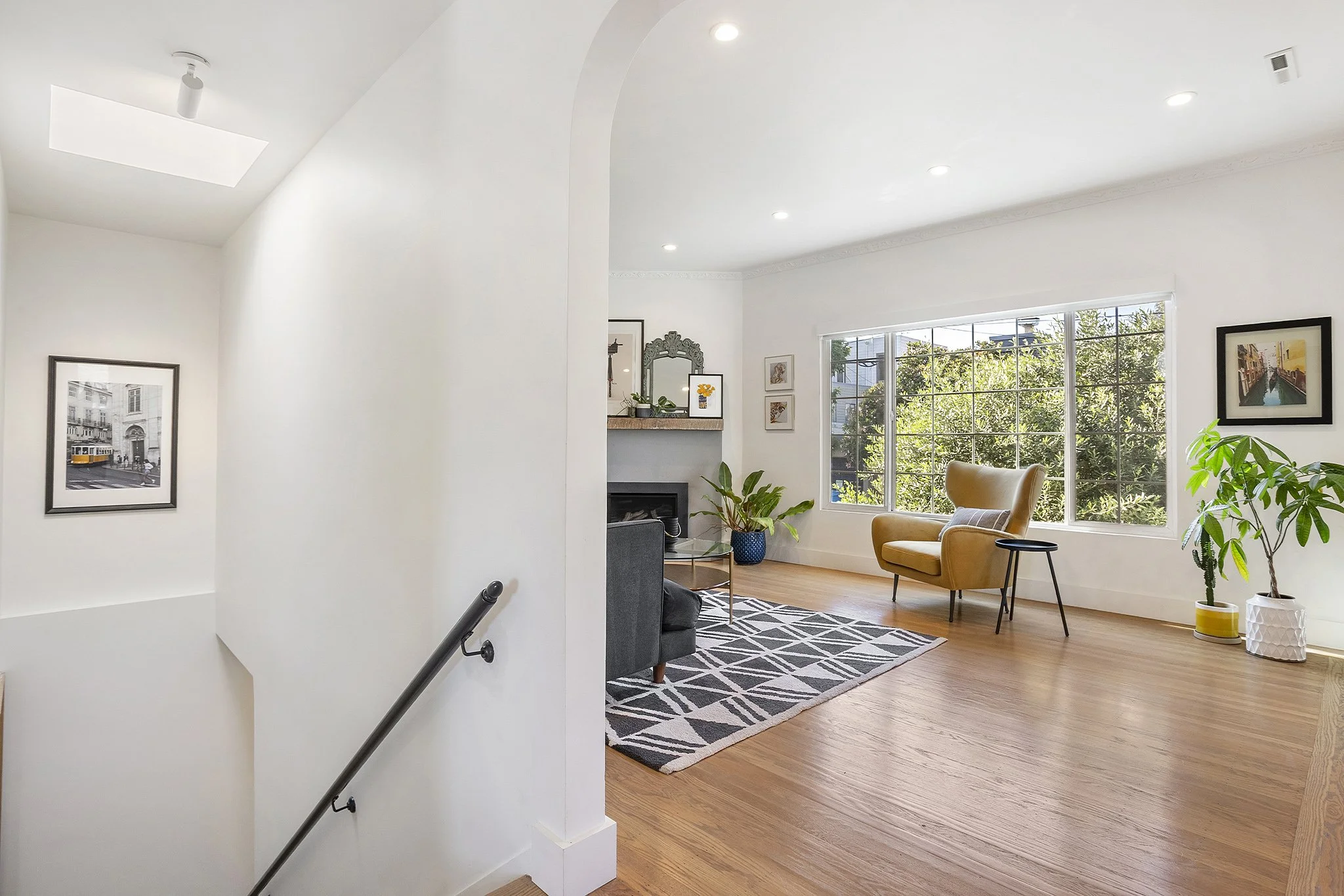 Bright living room with large window, yellow armchair, black patterned rug, green plants, and artwork on white walls.