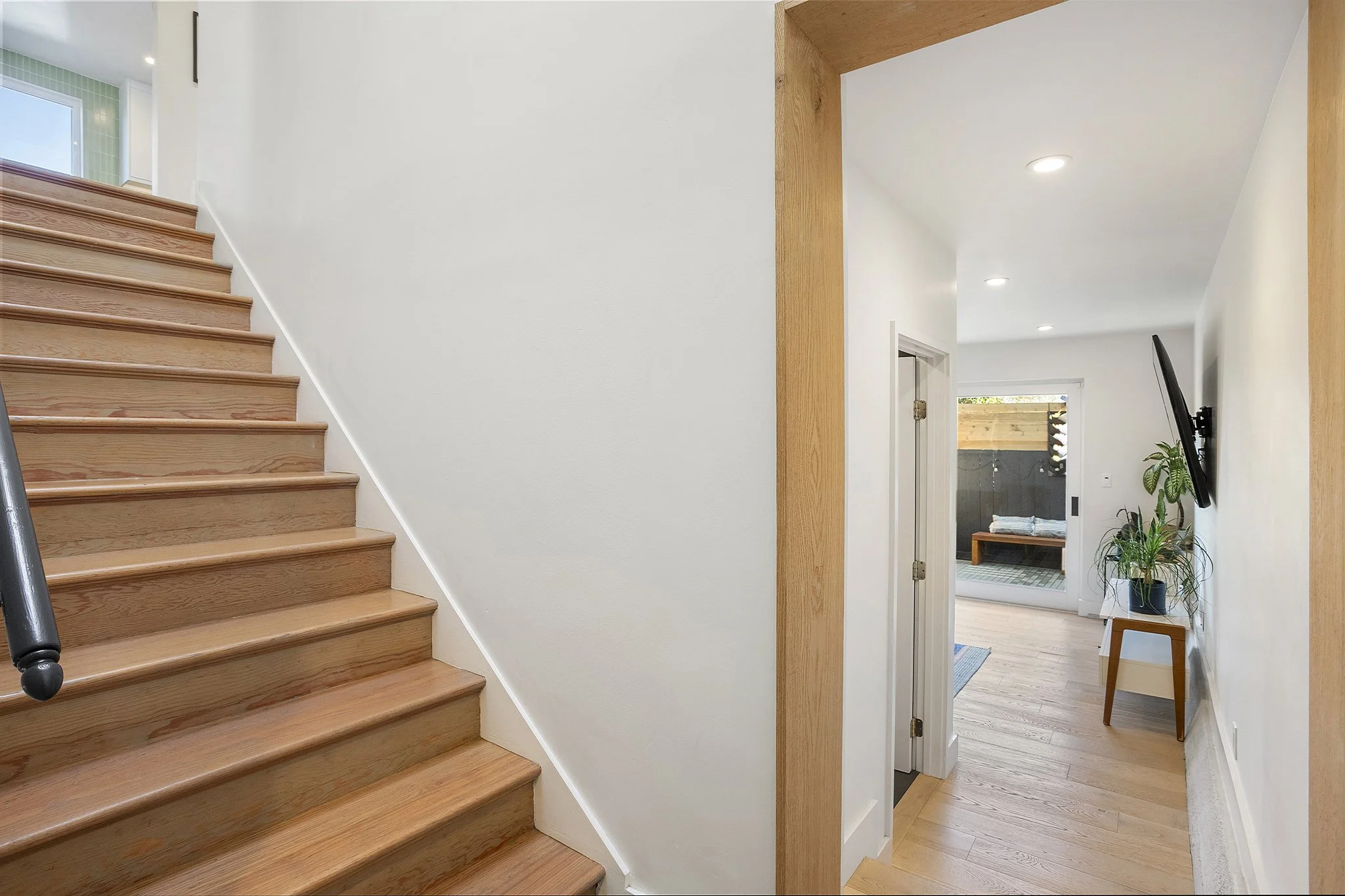 Interior view of a modern house with wooden stairs on the left, leading upstairs and a hallway on the right with a TV mounted on the wall, a window, and a potted plant on a small table.