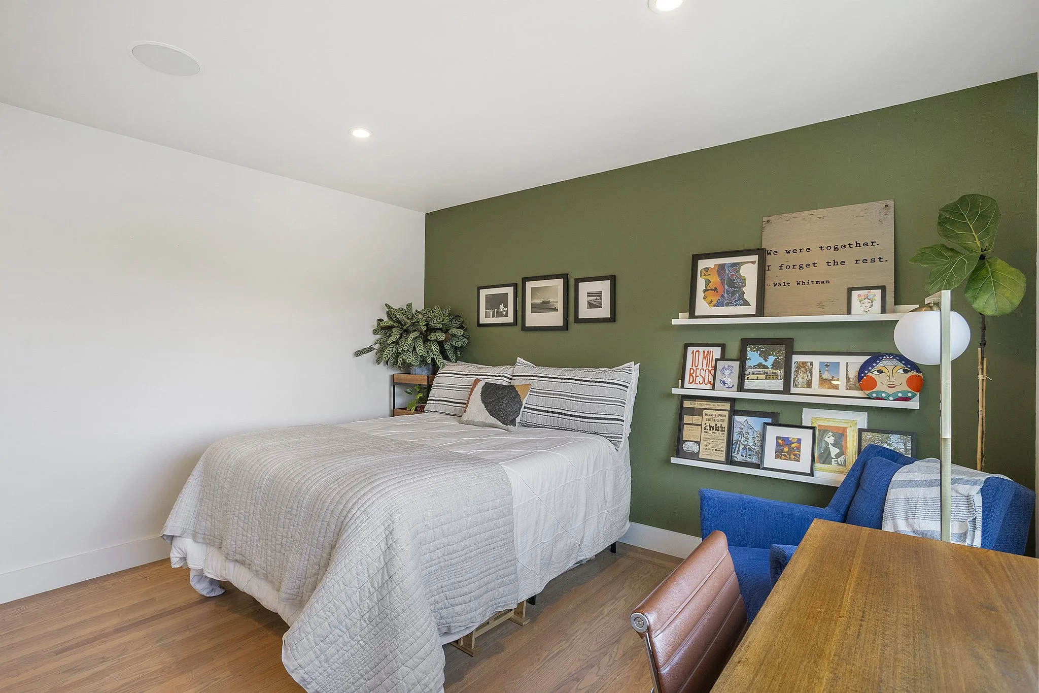 A bedroom with a bed against a green accent wall, decorated with framed art and a potted plant. There is a blue armchair, a wooden table, and a white floor lamp with a round shade.