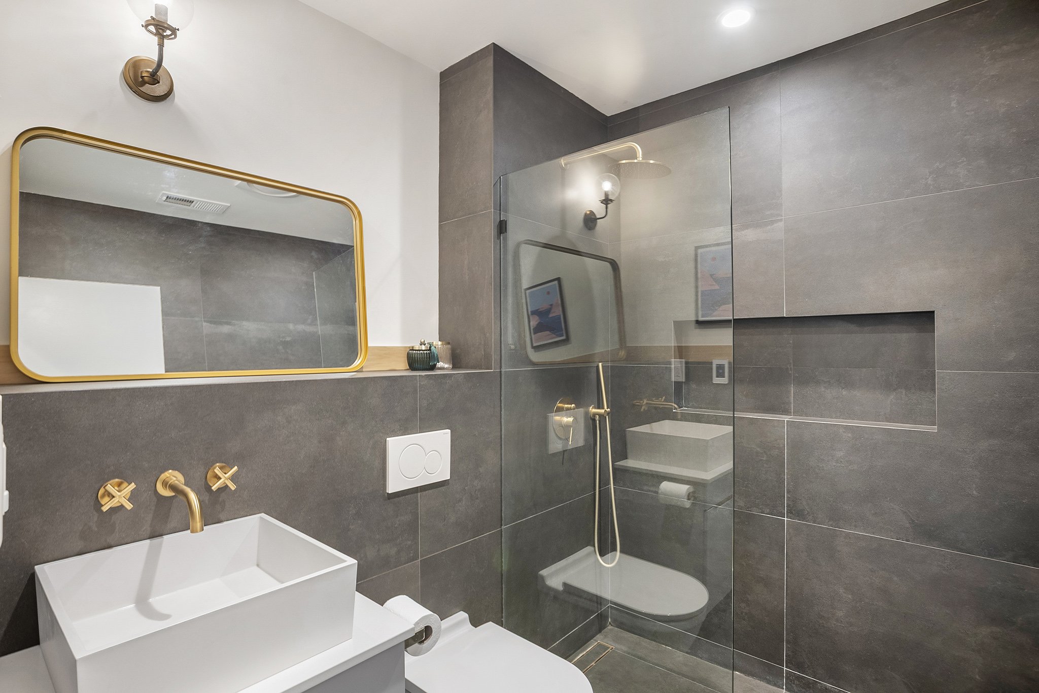 Modern bathroom with gray tiles, white sink with gold faucet, large mirror with gold trim, shower with glass partition, and wall-mounted light fixtures.