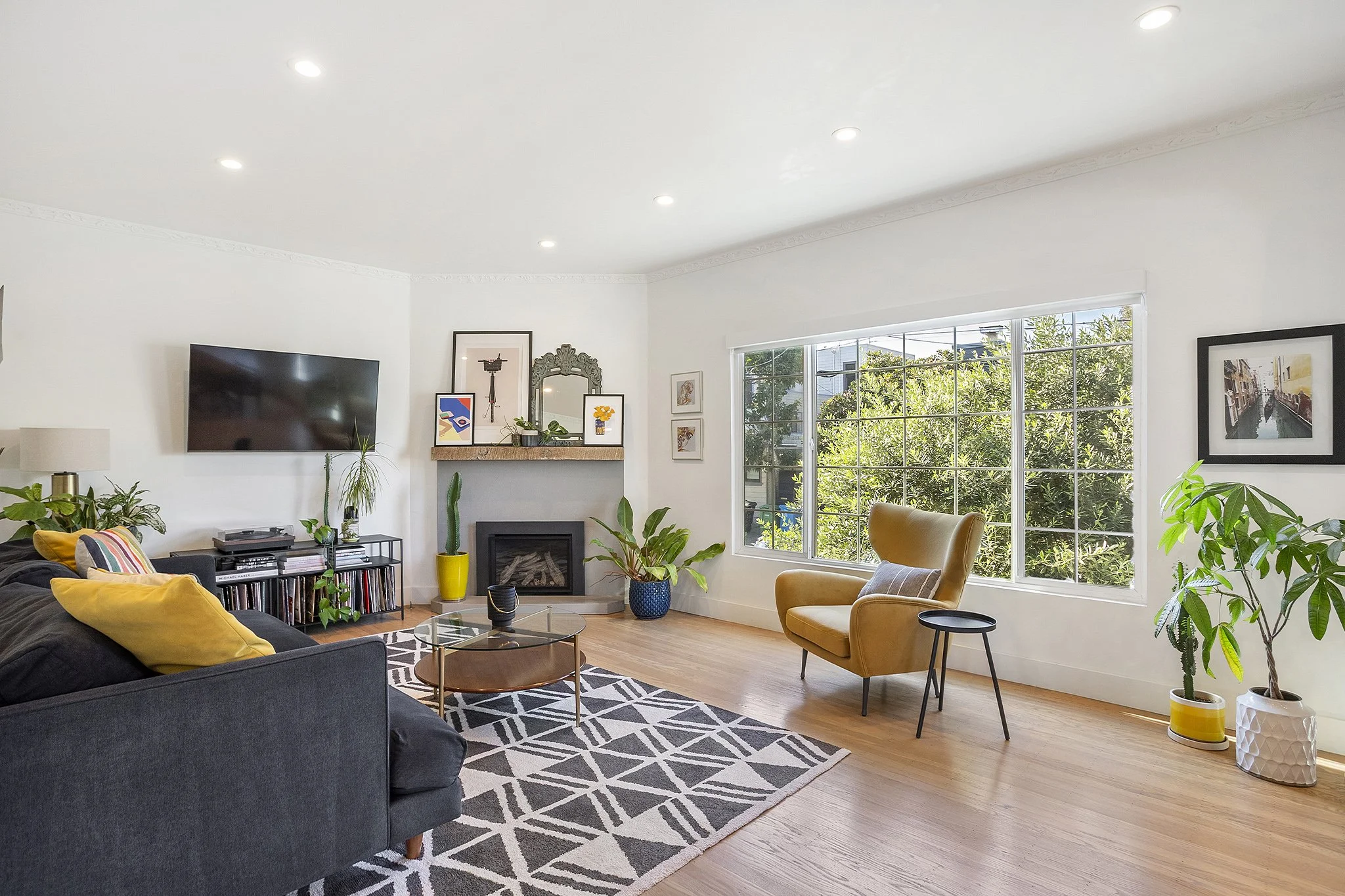 Bright living room with large window, black sofa with yellow pillows, yellow armchair, striped rug, glass coffee table, potted plants, wall art, and a television on white walls.