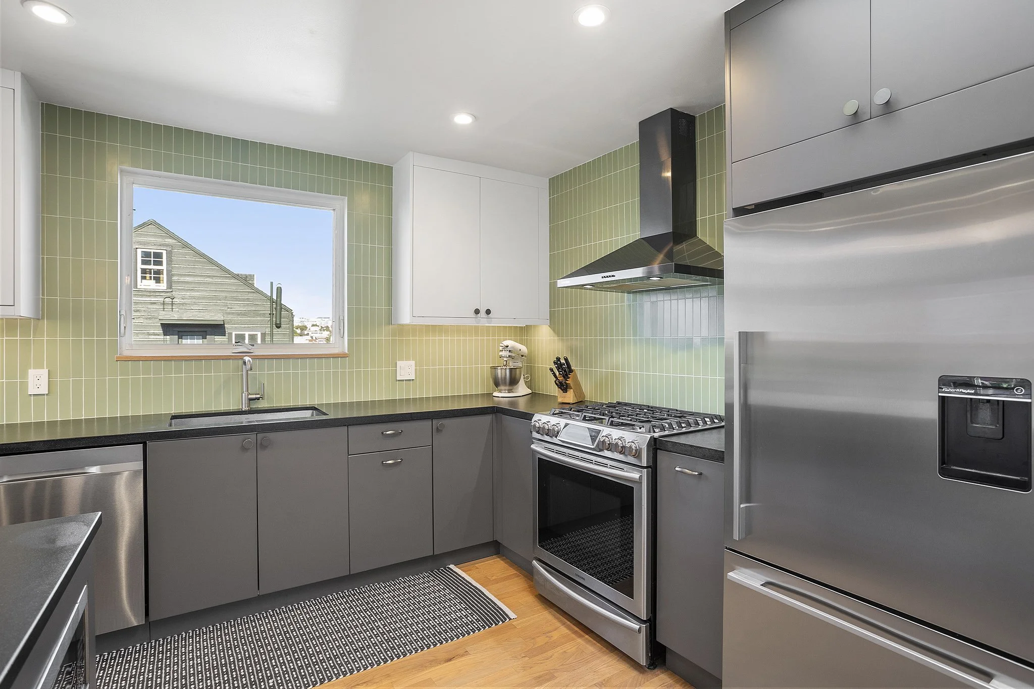 Modern kitchen with gray cabinets, stainless steel refrigerator, oven, range hood, window, black countertop, green tiled backsplash, and hardwood floor.