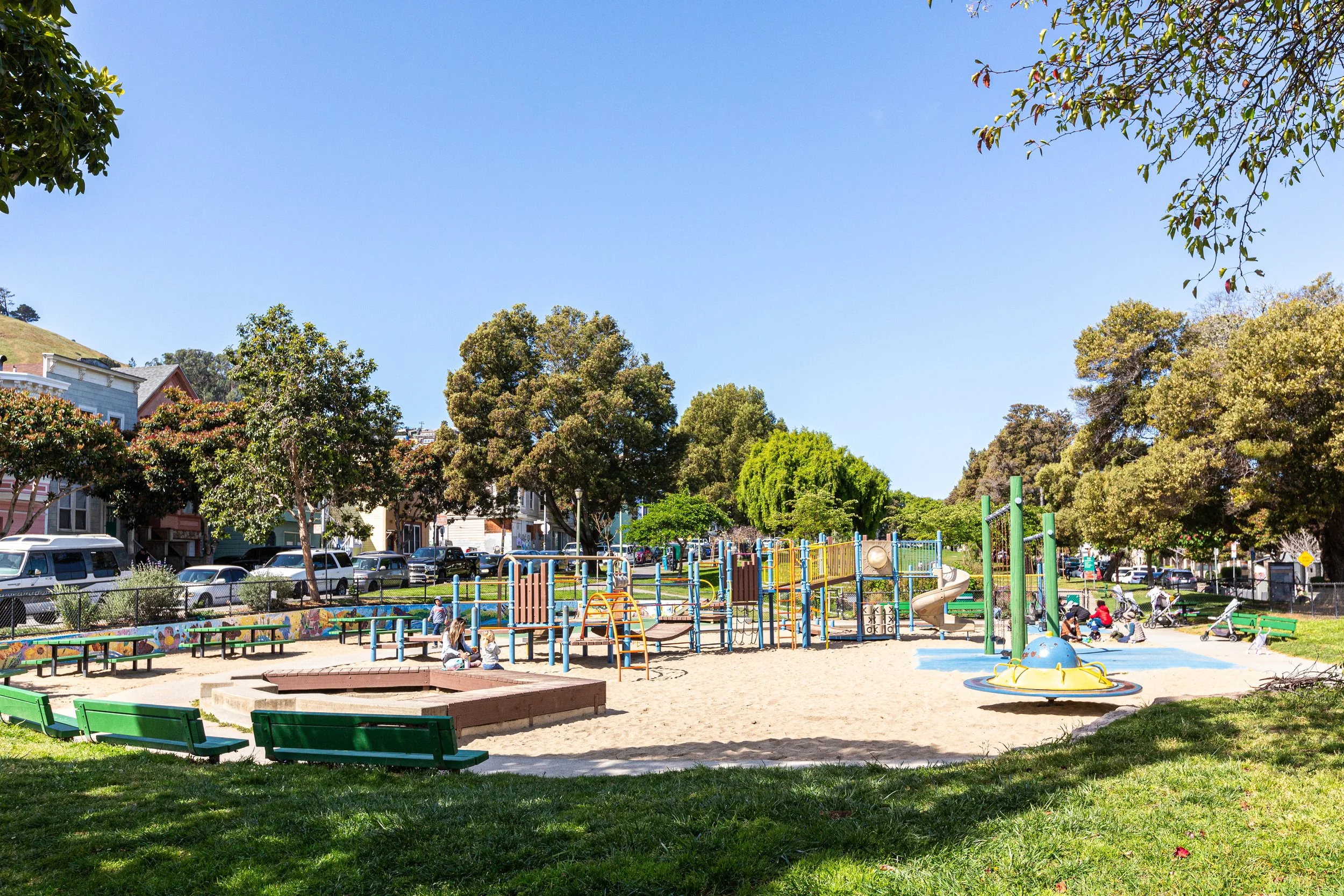Children playing on playground equipment, including slides and climbing structures, in a park with trees, grass, and benches, under a clear blue sky.