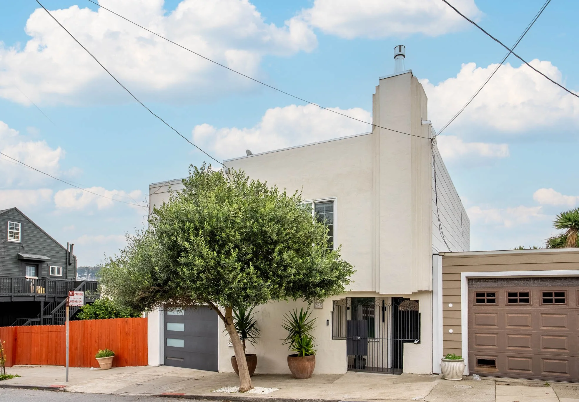 Front view of modern white residential building with trees and potted plants, garage door, and a sidewalk.