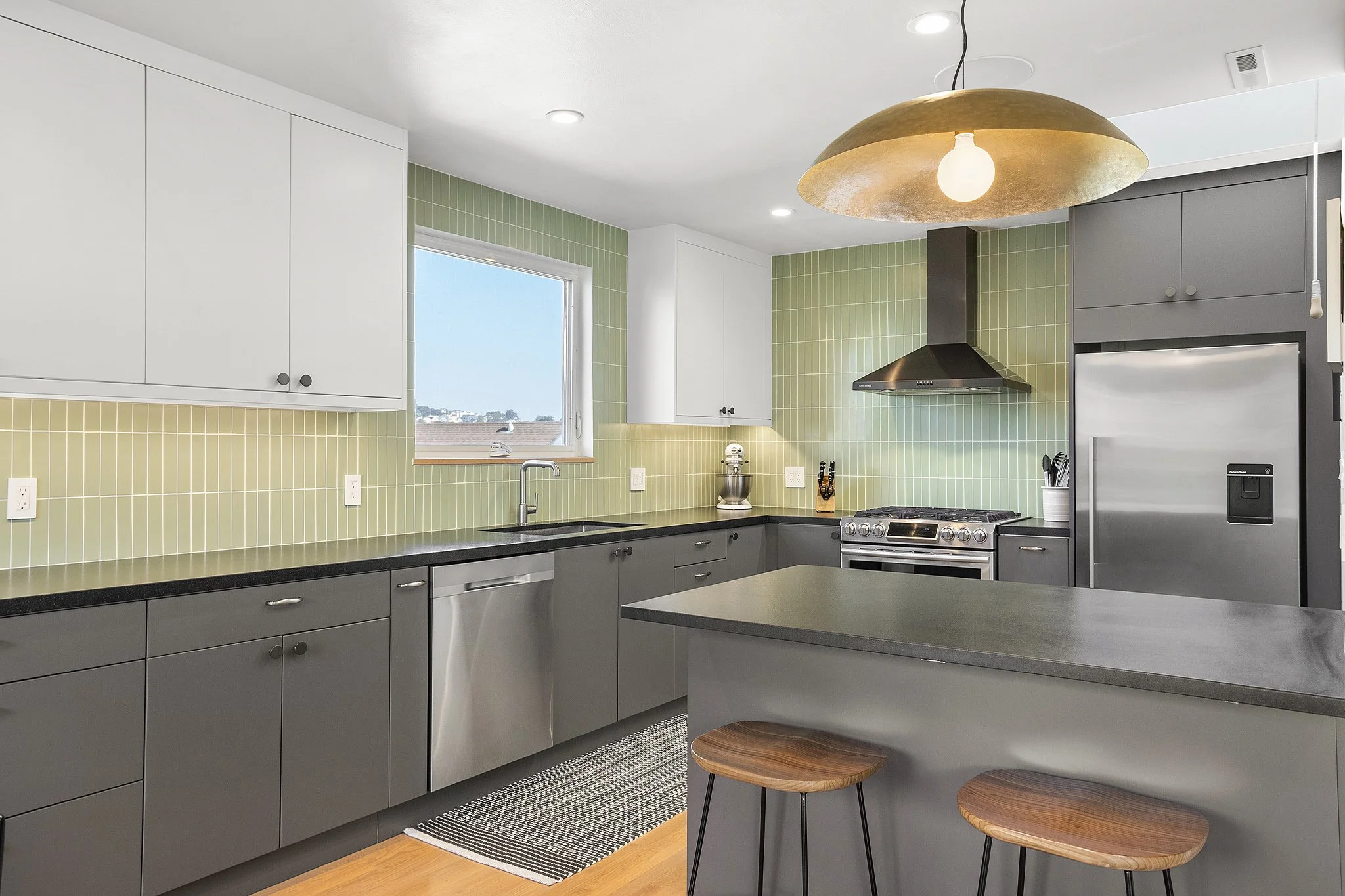 Modern kitchen with grey cabinets, a black countertop, green tile backsplash, stainless steel appliances, a window, and a gold pendant light