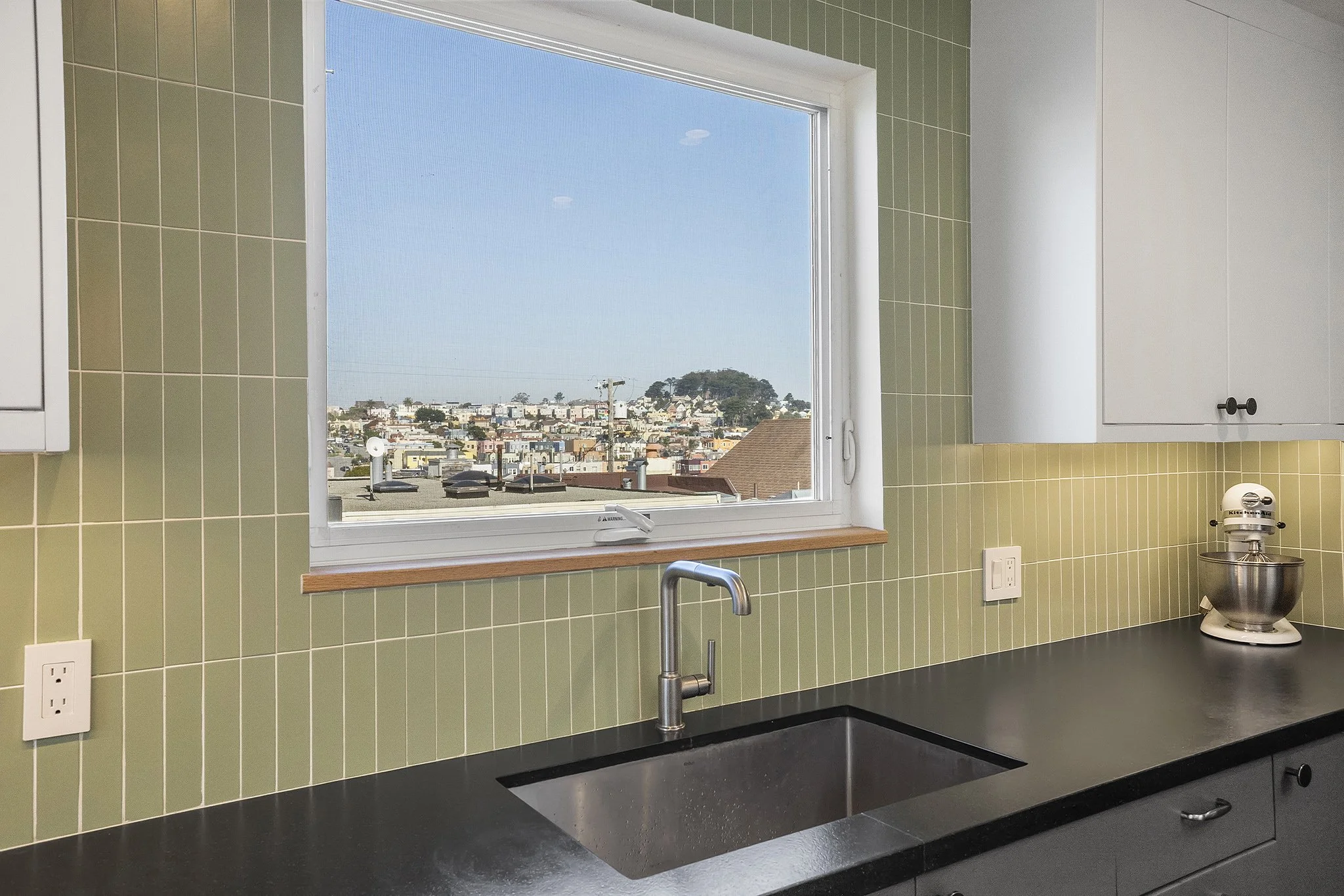 Kitchen with green tiled wall, black countertop, stainless steel sink, white cabinets, and a window showing a cityscape and hills in the background.