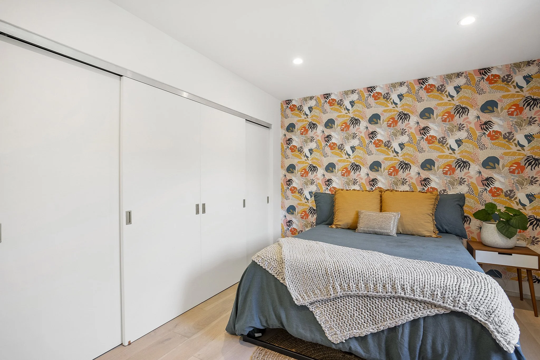 A bedroom with a bed covered in blue bedding and layered with beige and gray pillows, a white chunky knit blanket, next to a side table with a potted plant, and a colorful patterned accent wall behind the bed.