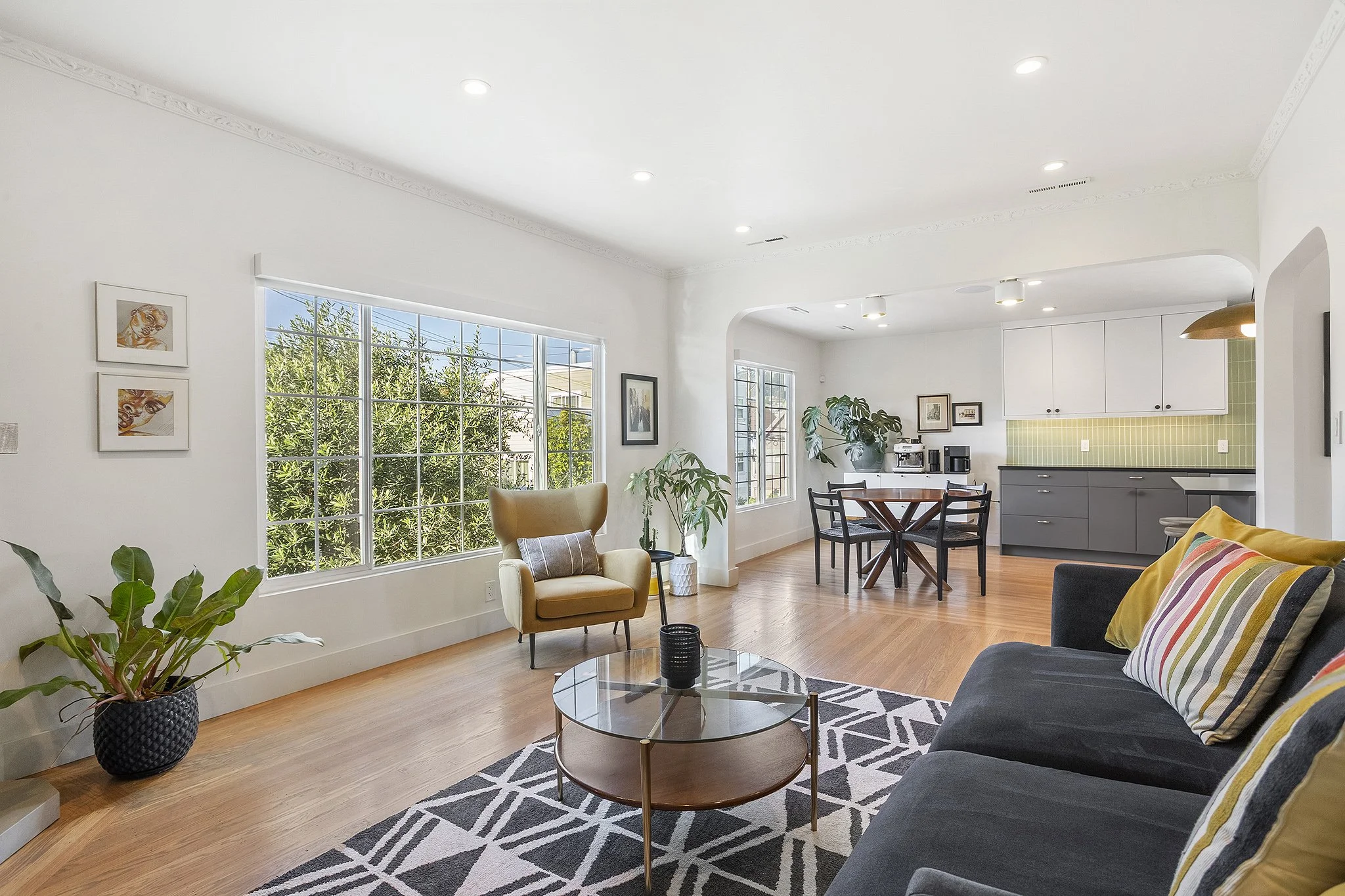 Bright open living room with large window, wooden floors, gray sofa with yellow pillows, beige armchair, round coffee table, indoor plants, and a view into the kitchen with white cabinets and green backsplash.
