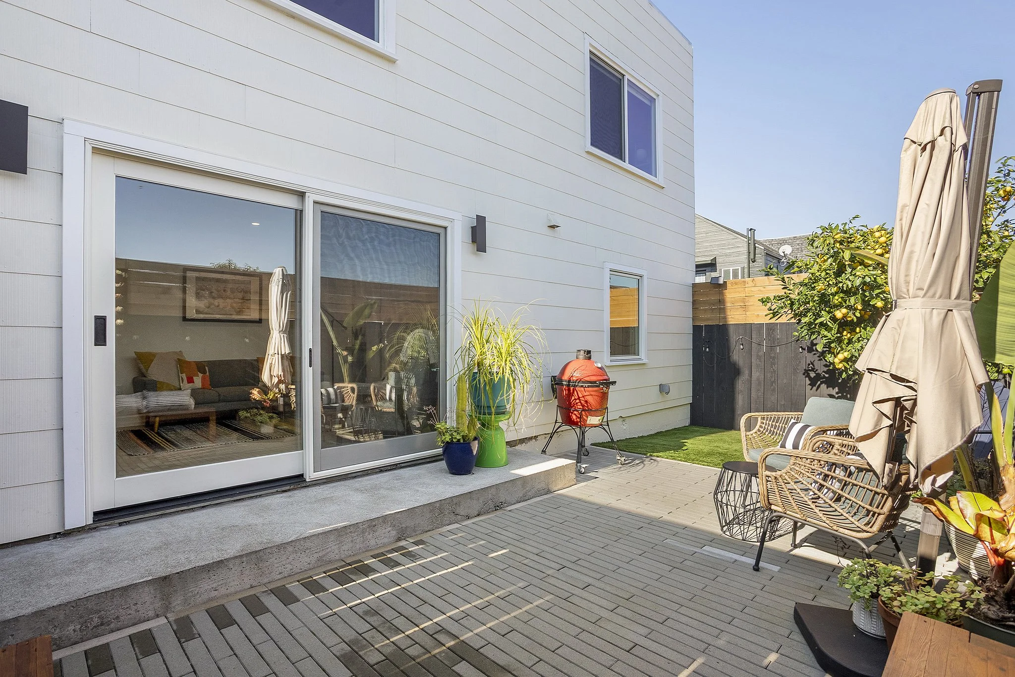 Backyard patio with potted plants, a barbecue grill, outdoor chairs, a large patio umbrella, and a sliding glass door leading into a house with a white exterior and a view of the interior living room.