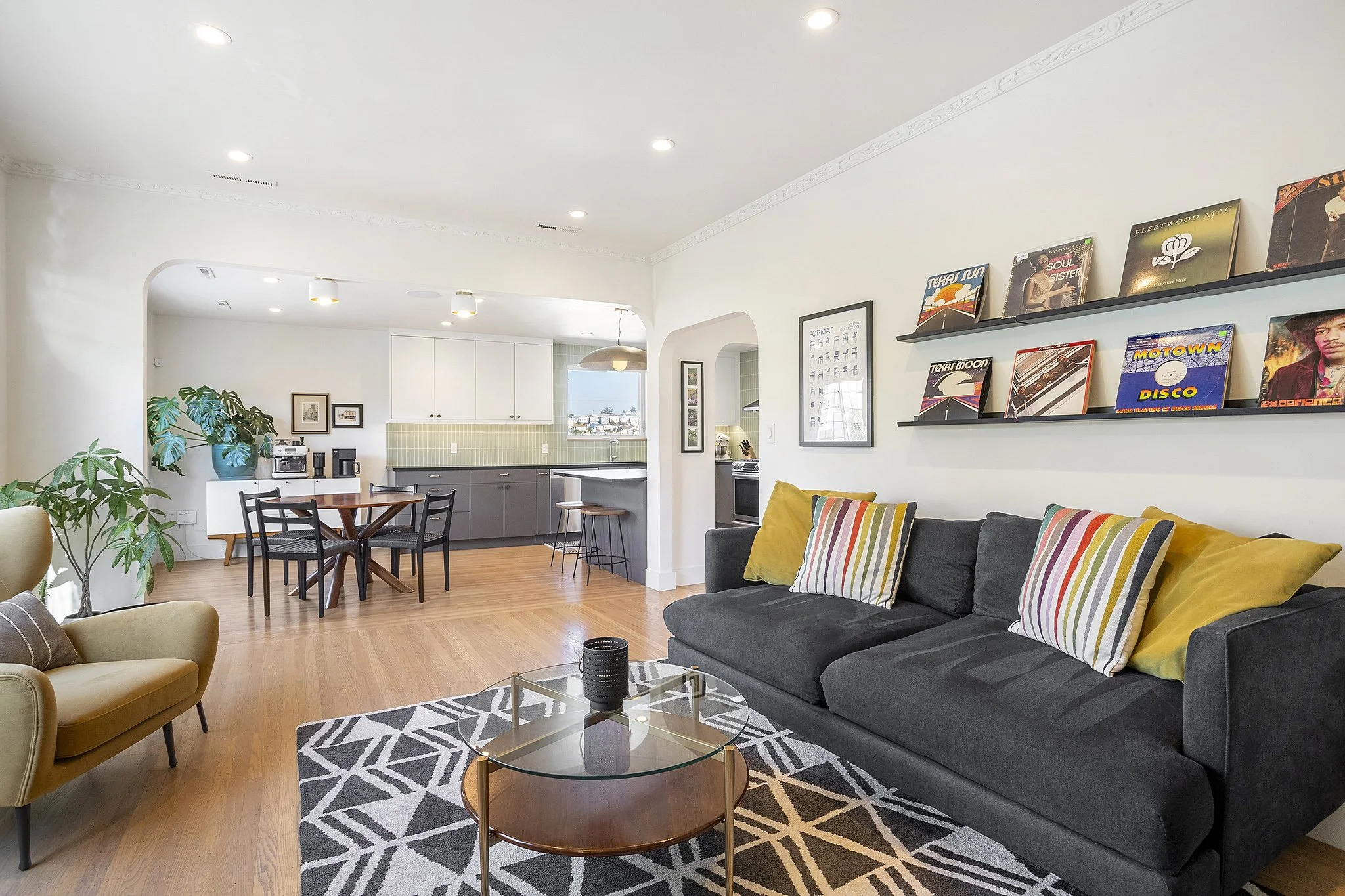 Living room with black sofa, yellow and striped pillows, glass coffee table, geometric rug, and a bookshelf with vinyl records. Open kitchen and dining area with a round table, black chairs, and bar stools.