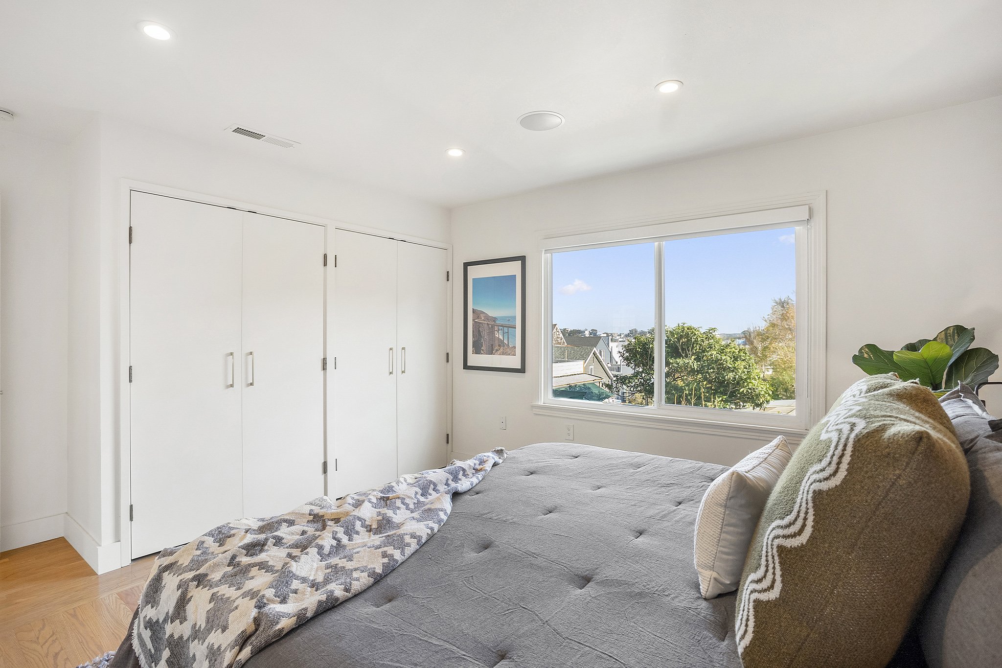 Bright bedroom with large window, white walls, and closet doors. Bed partially made with a gray sheet, an unmade blanket, and pillows, featuring a potted plant nearby and framed art on the wall.
