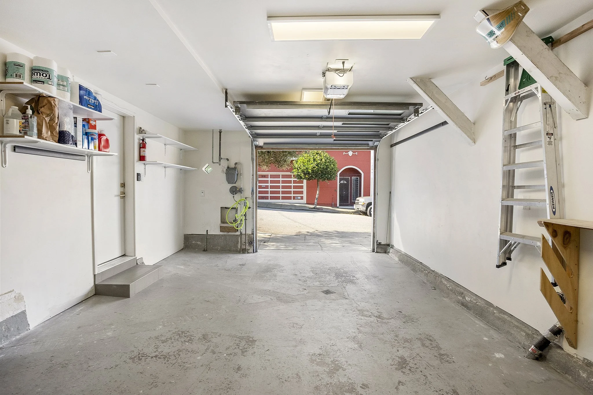 View of a clean, empty residential garage with white walls, shelves on the left holding containers and supplies, an open garage door showing the street with a tree and red building outside, a ladder on the right wall, and a ceiling-mounted garage doo