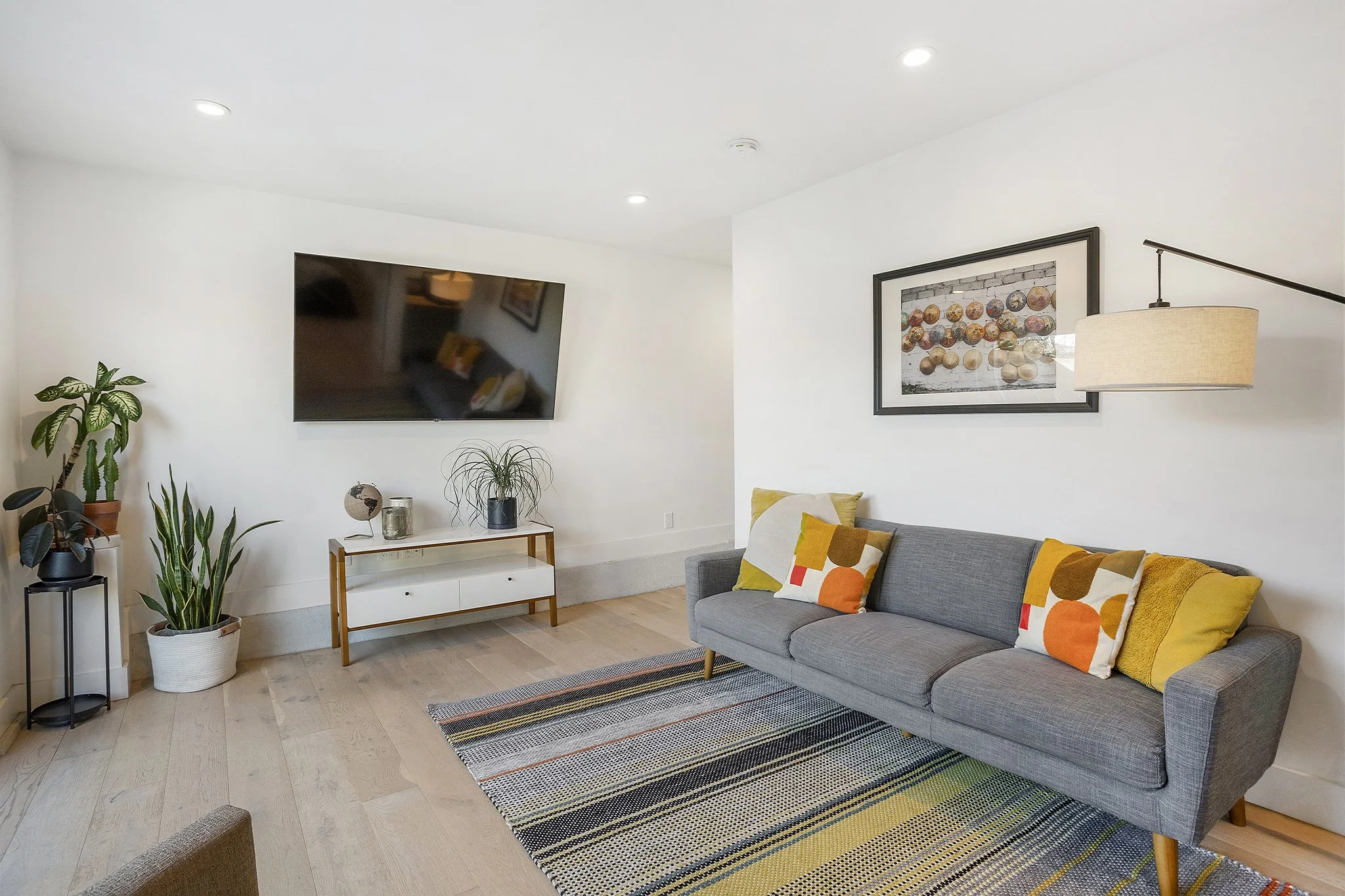 Living room with a gray sofa, colorful pillows, wall art, a mounted flat-screen TV, and potted plants.