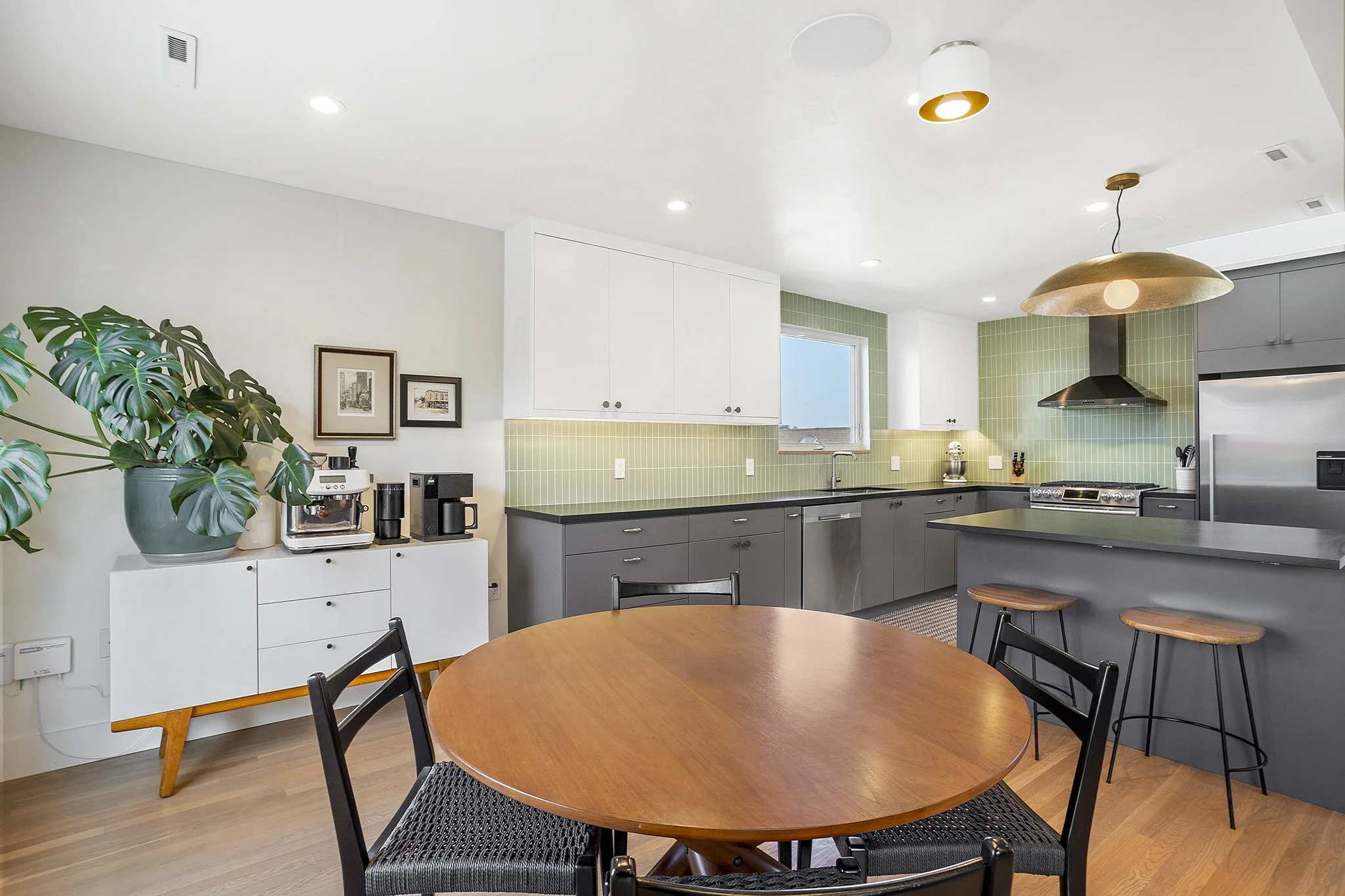 Modern kitchen interior with gray cabinets, white upper cabinets, green tile backsplash, stainless steel appliances, wooden dining table with black chairs, large potted plant, and pendant lighting.