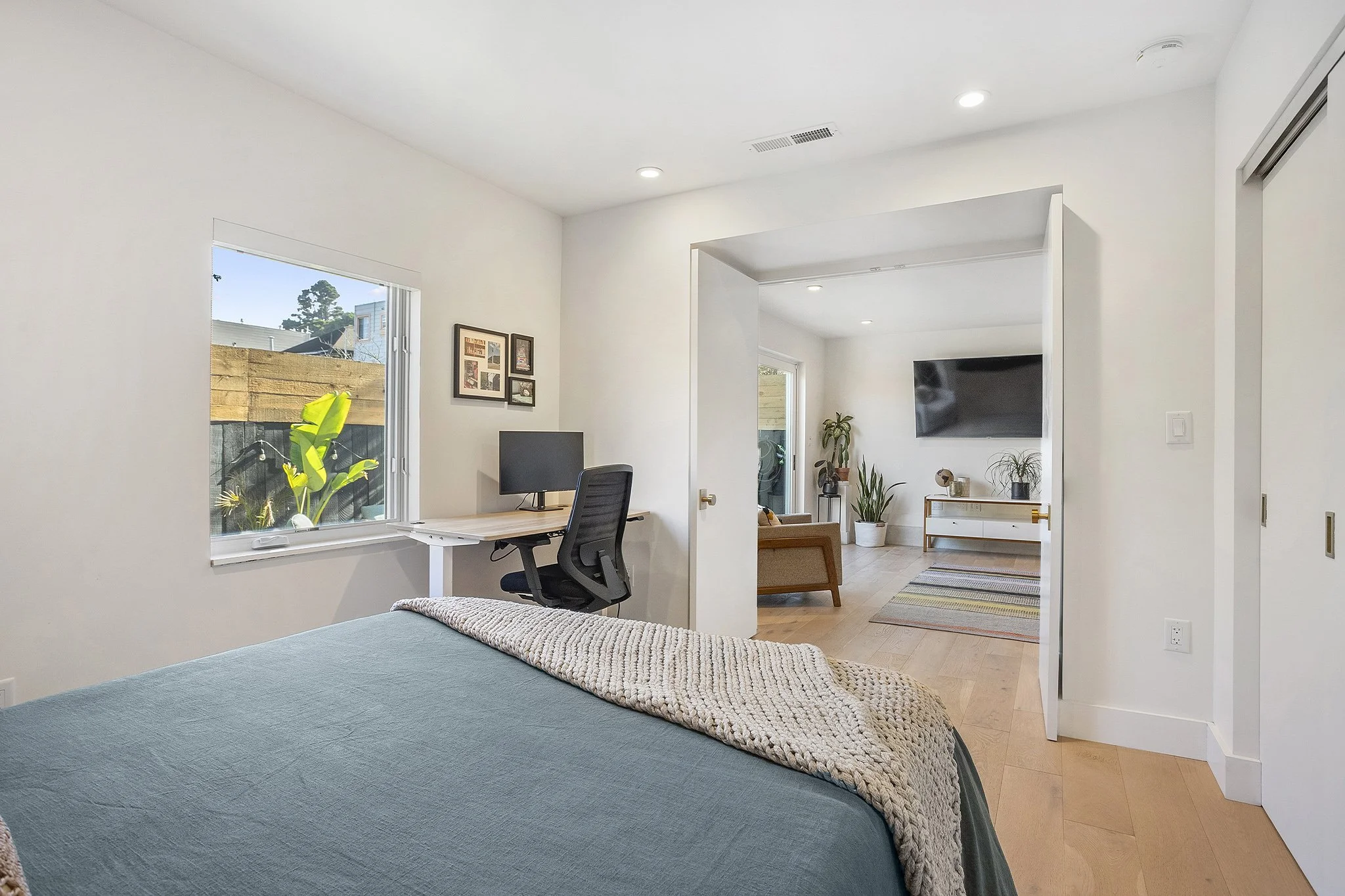 A bedroom with a bed, small desk with a computer, and open door leading to a living room with a TV, plants, and a rug.