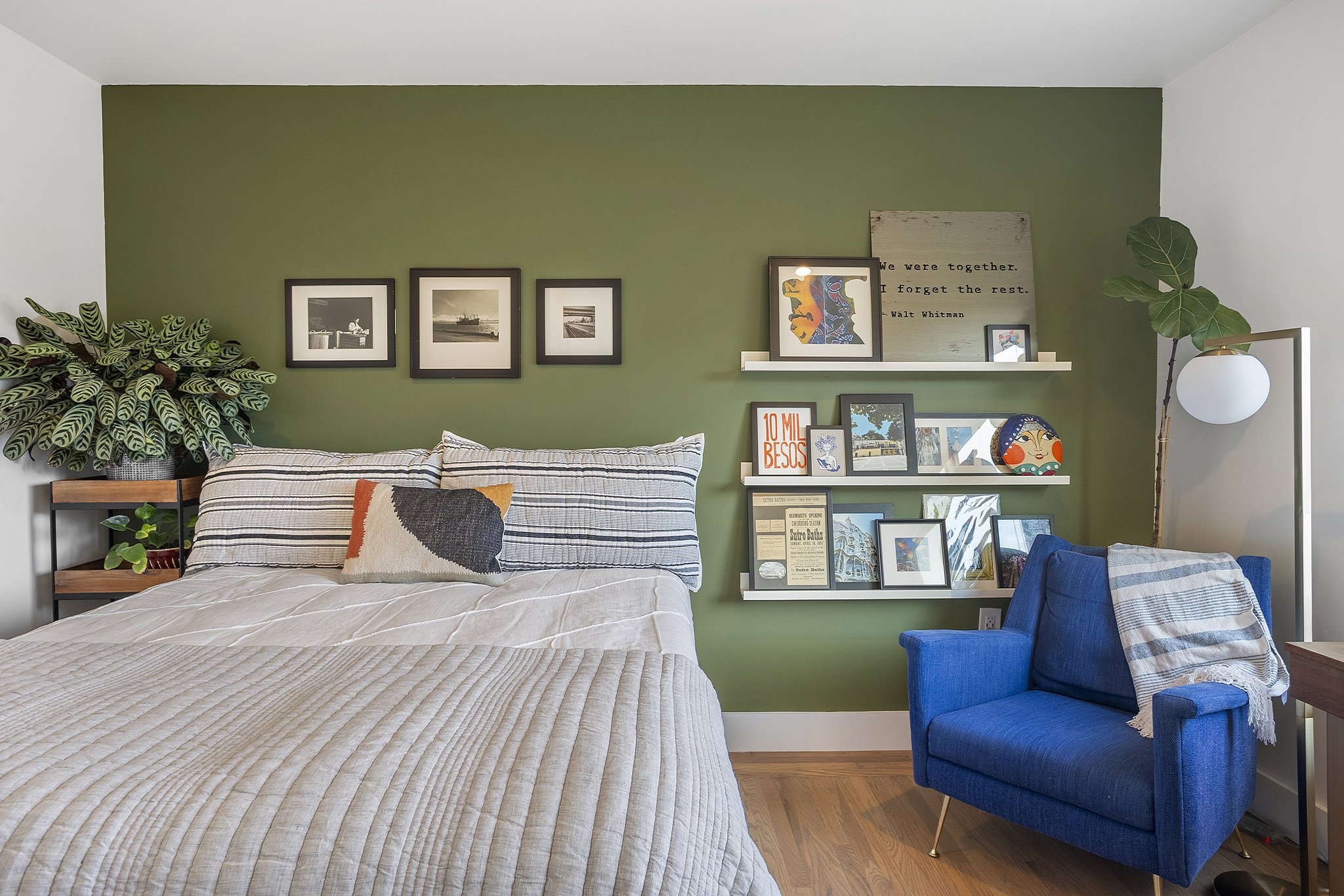 Bedroom with green accent wall, framed black and white photos above bed, patterned throw pillow on bed, blue armchair with blanket, white shelving with various framed pictures and decorations, large indoor plant on side table, standing lamp with roun