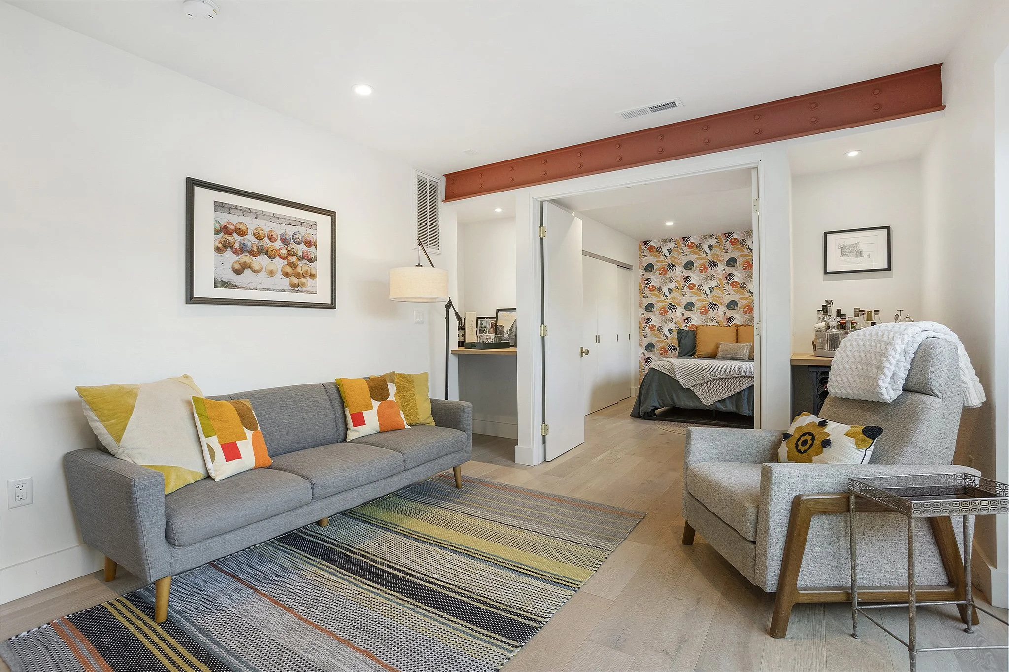 Living room with a gray sofa, colorful throw pillows, a gray armchair with a small pillow and blanket, a striped rug, and a doorway leading to a bedroom with patterned wallpaper.
