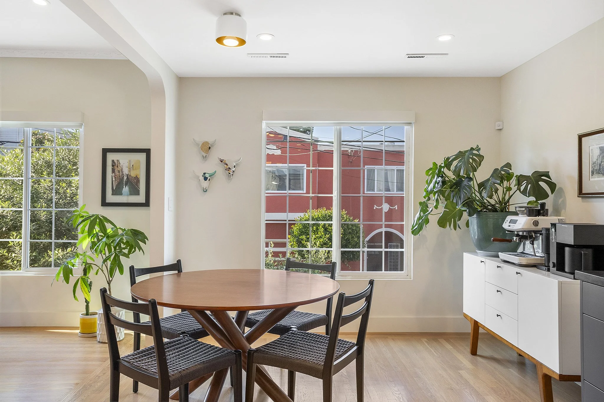 Dining area with a round wooden table, four black chairs, large window with a view of a red building and greenery, potted plants, wall decorations, and a cabinet with a coffee machine and a potted plant.