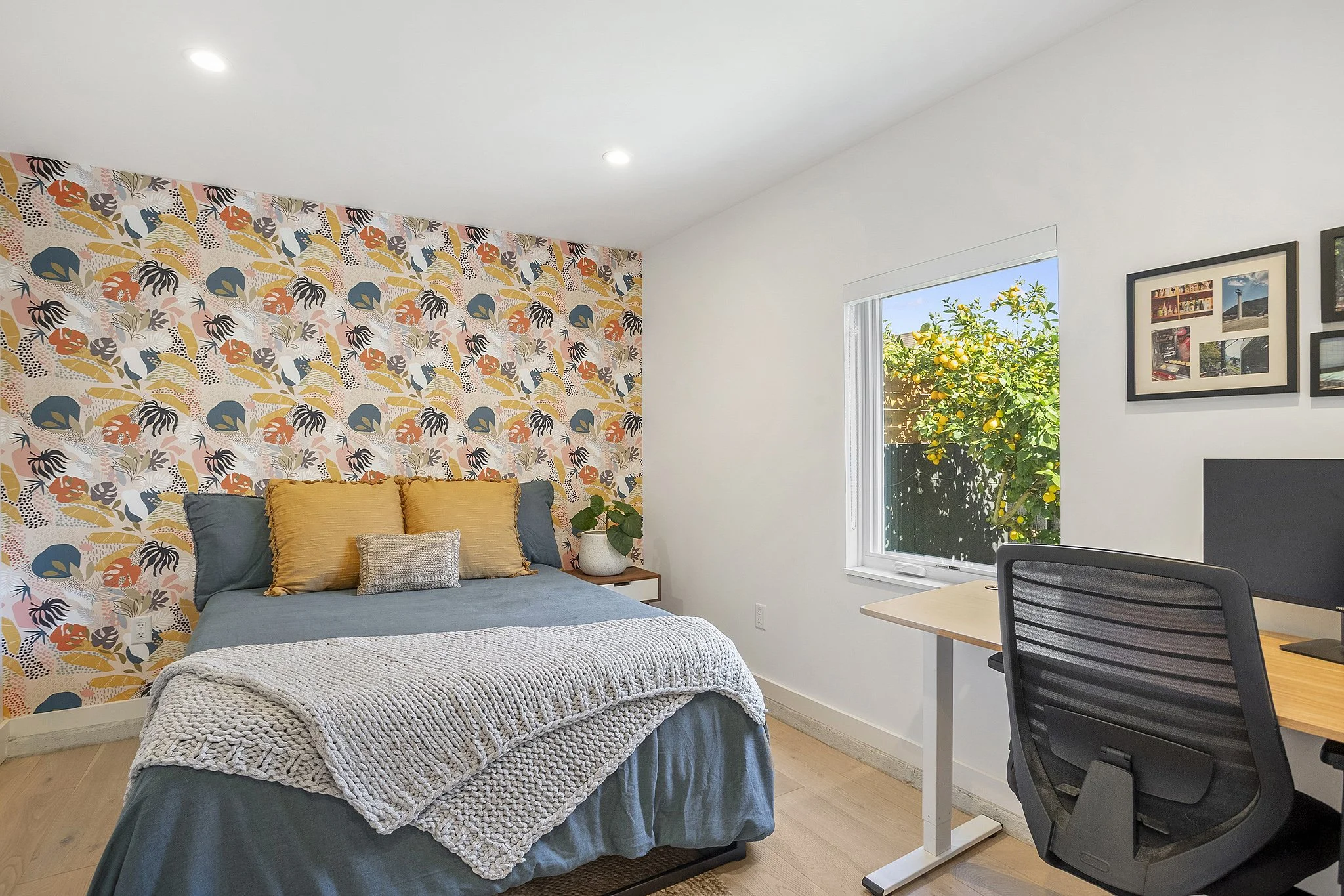 Bedroom with a bed covered in blue and white bedding, colorful floral and animal patterned wallpaper behind the bed, a bedside table with a plant, a window showing lemons on a tree outside, and a desk with a computer and framed photos on the wall.