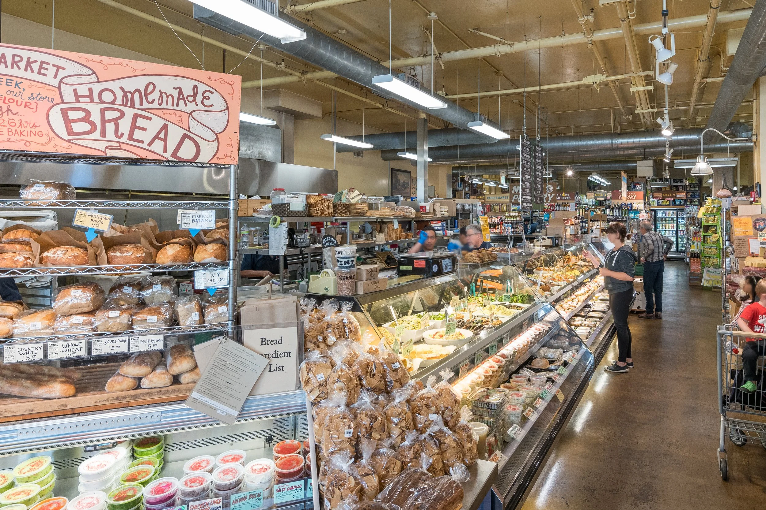 Interior of a grocery store focusing on the bakery section with fresh bread and baked goods on display, customers shopping, and store signage.