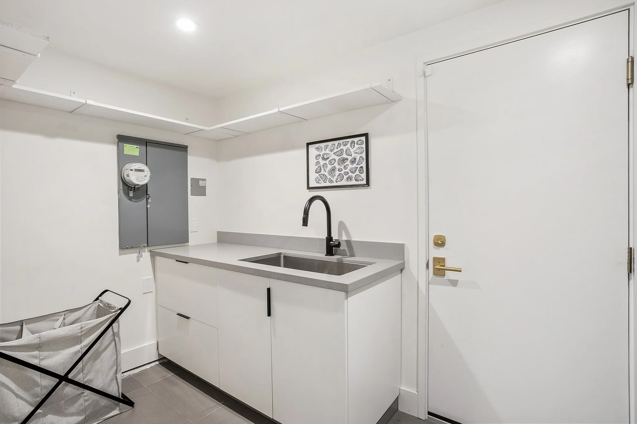 Minimalist laundry or utility room with white walls, gray countertop, black faucet, white cabinets, framed artwork of seashells, ceiling light, and a laundry hamper.