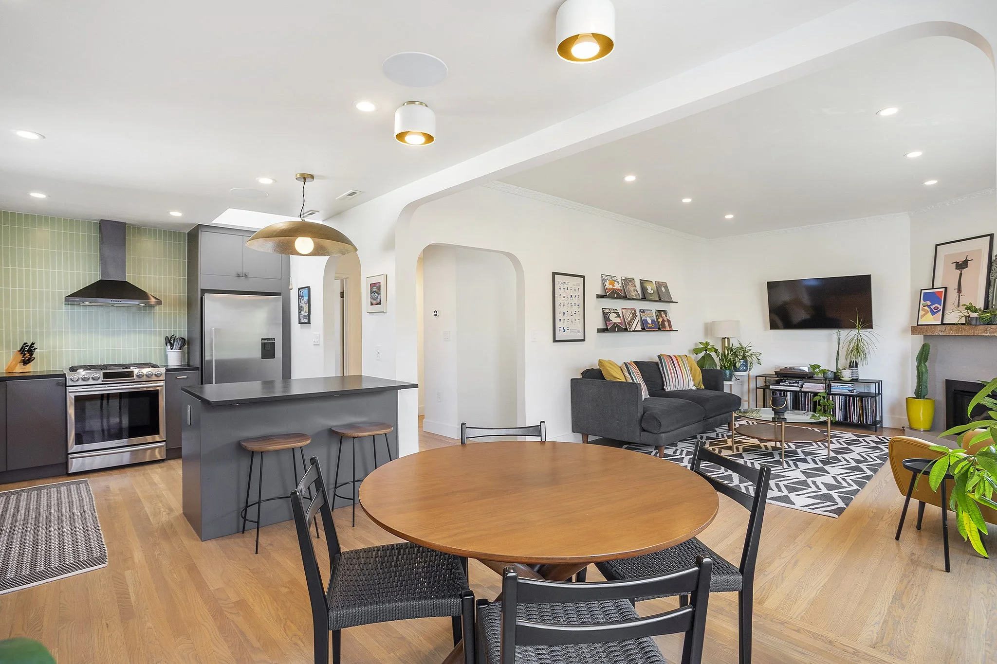 Open-concept living room and kitchen with wooden flooring, gray cabinets, black appliances, a round wooden dining table with black chairs, and a sitting area with a gray sofa, wall-mounted TV, and various decorative plants.