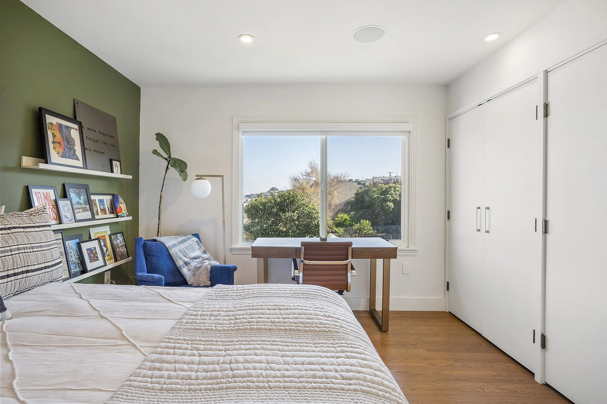 A bedroom with a window showing trees and buildings outside, a bed with a textured cream-colored blanket, a green accent wall with framed pictures and a white shelf, a blue chair with a plaid throw, a wooden desk near the window, and white closet doo