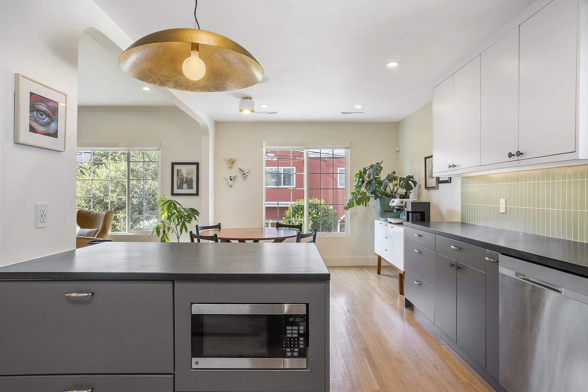 Modern kitchen with gray cabinets, black countertop, white upper cabinets, microwave, dishwasher, and green tile backsplash. Dining area with wooden table, black chairs, and potted plants near windows with sunlight.