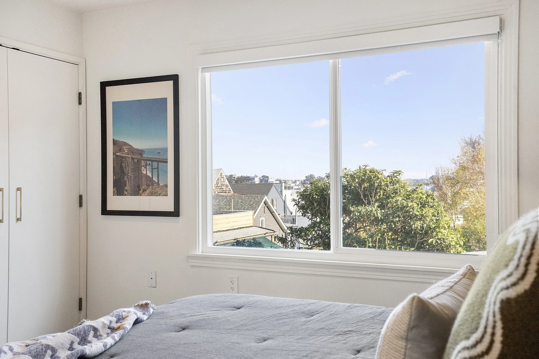 View of a bedroom with a large window showing a neighborhood and blue sky, a framed photo of a coastline on the wall, and a bed with gray bedding and pillows.