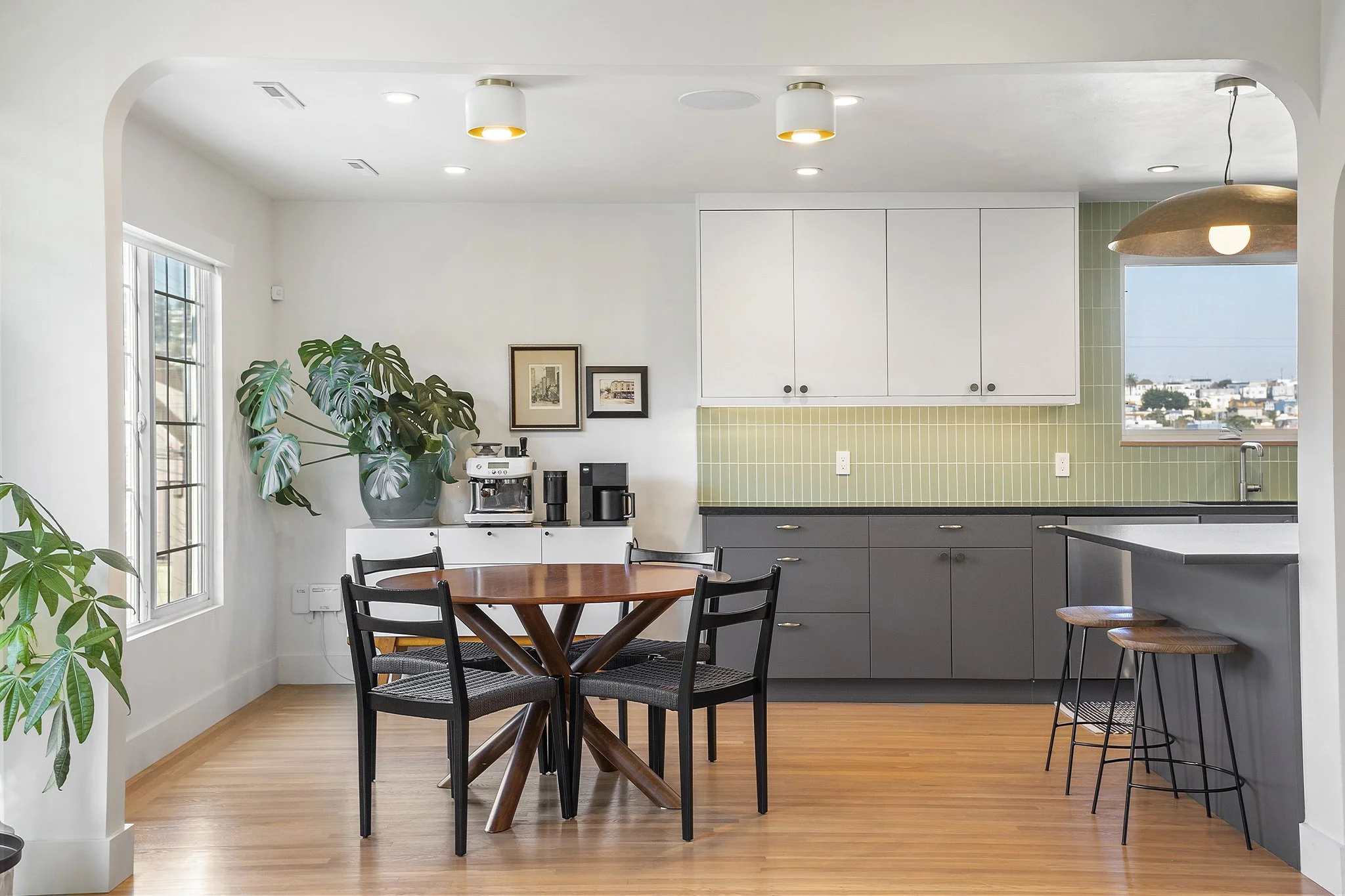 Modern kitchen with white and green cabinets, a round wooden dining table with black chairs, large windows, and a coffee station.