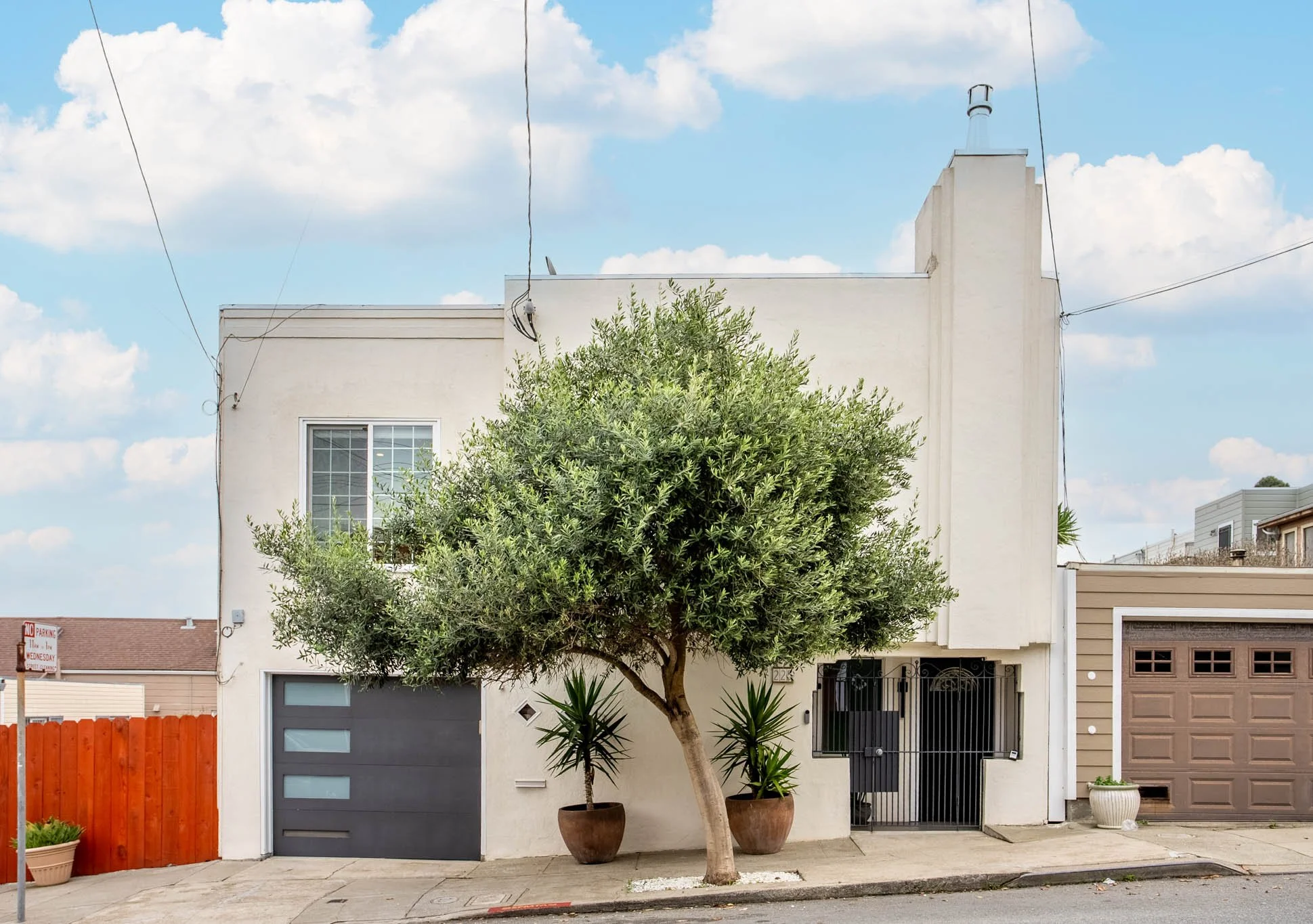 White multi-story house with a large tree in front, two potted plants, a black door with a gate, and a brown garage door. Clear sky with clouds.
